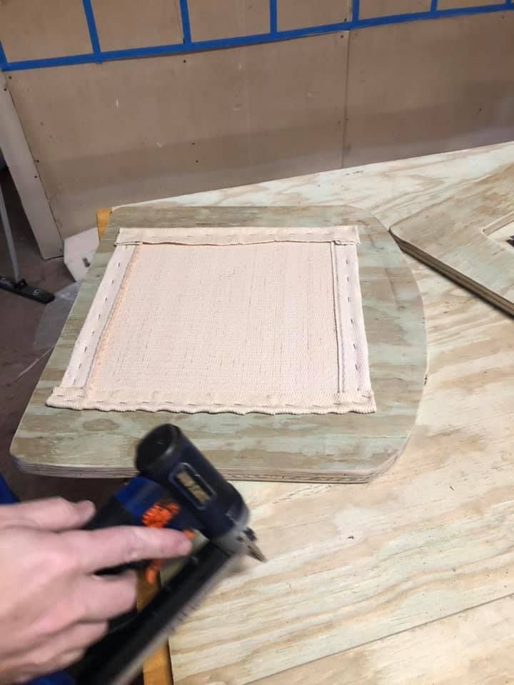 Person stapling fabric to a wooden chair seat. The fabric is light brown, and the wood is light green.