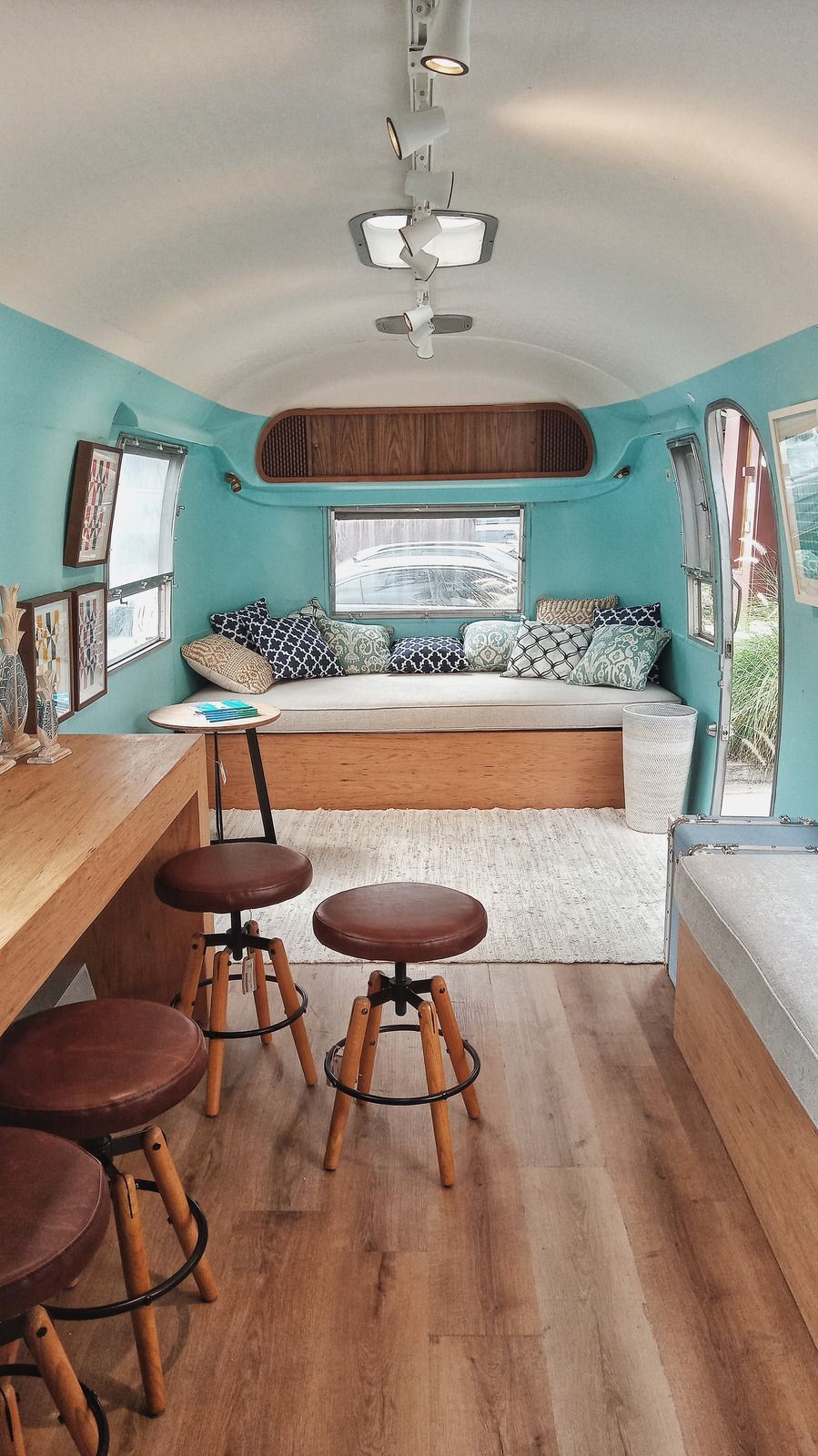 Interior of a teal-painted retro camper with a wooden bar, stools, and a bed area with pillows.