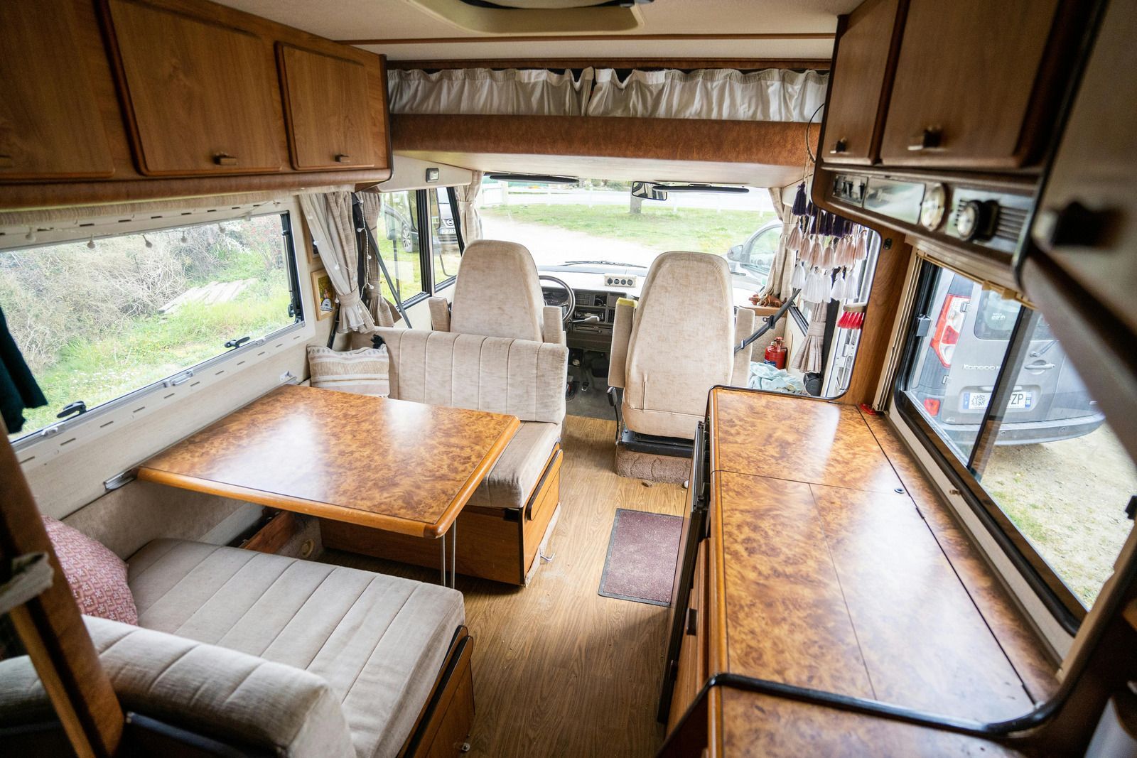 Interior view of a vintage RV with wooden cabinets, seating, table, and a view through the front windshield.