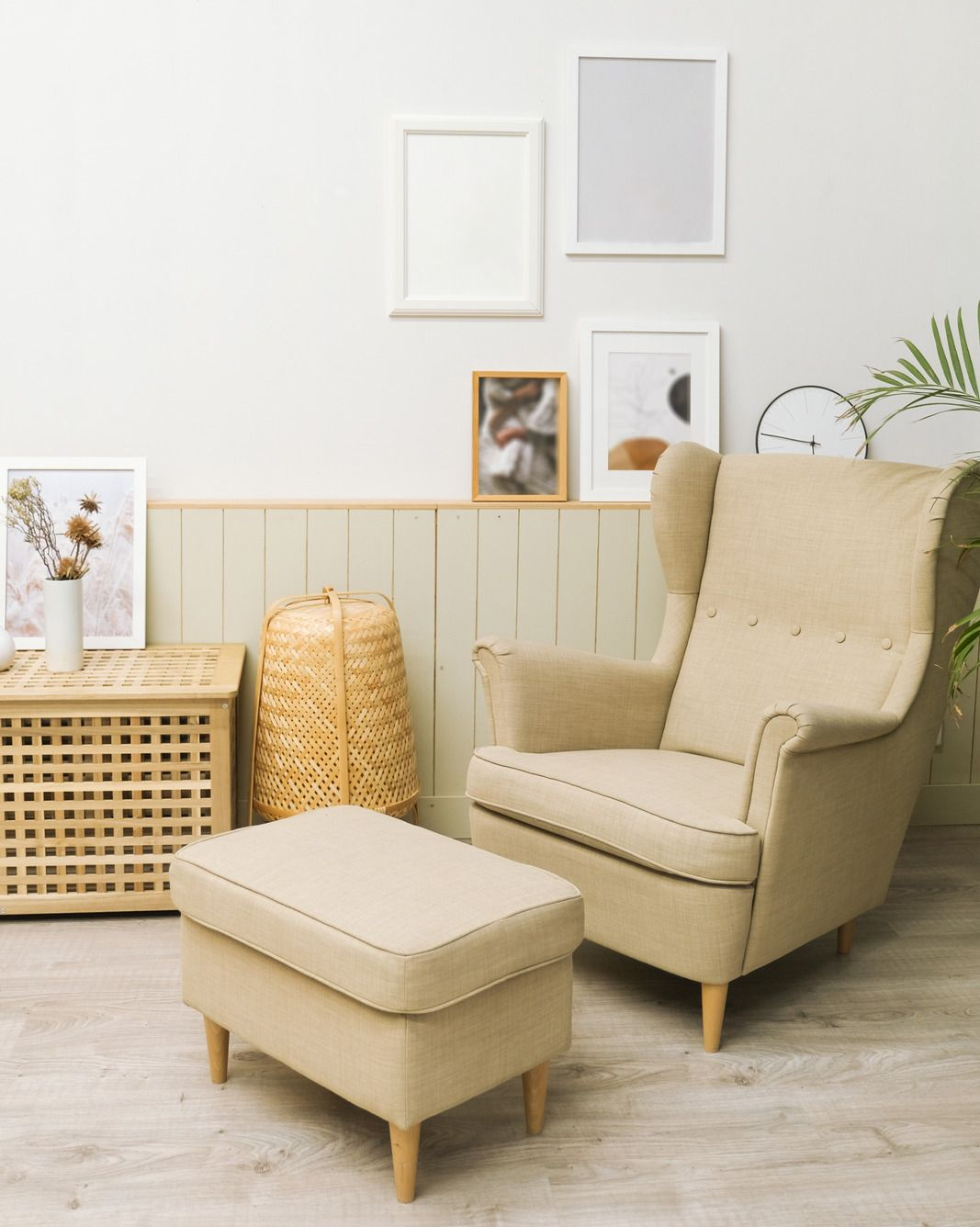 Beige armchair with ottoman in a room, next to a woven basket and wooden cabinet, frames on the wall.