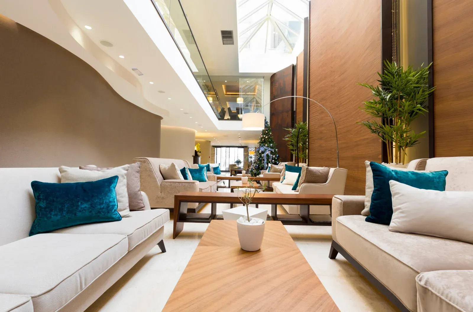 Lounge area with light-colored sofas, teal pillows, and wooden tables. Tall windows and plants add natural light.