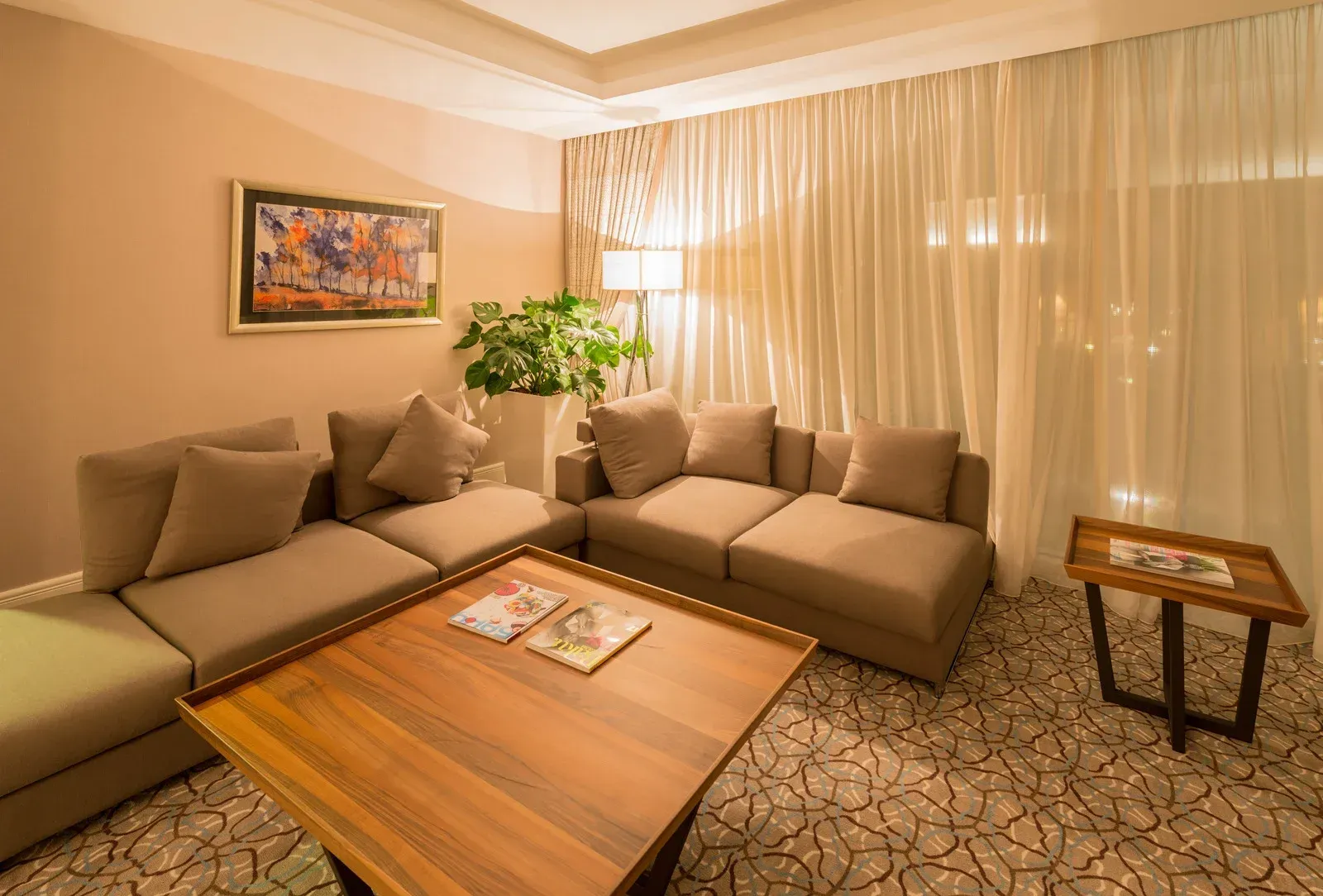 Living room with tan sectional sofa, coffee table, small side table, curtains, painting, and patterned carpet.