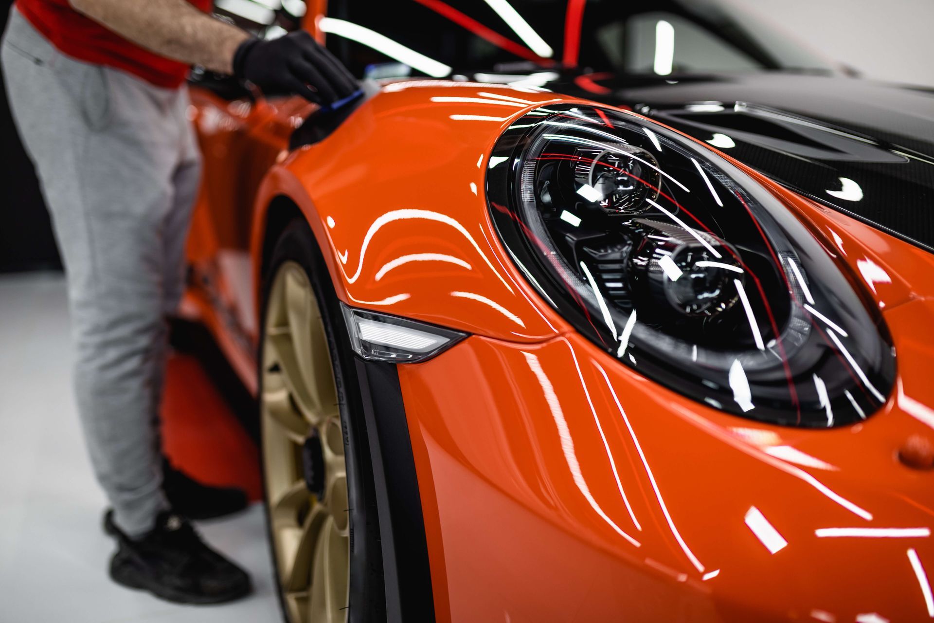 Person polishing a shiny orange sports car with black accents and gold wheels.