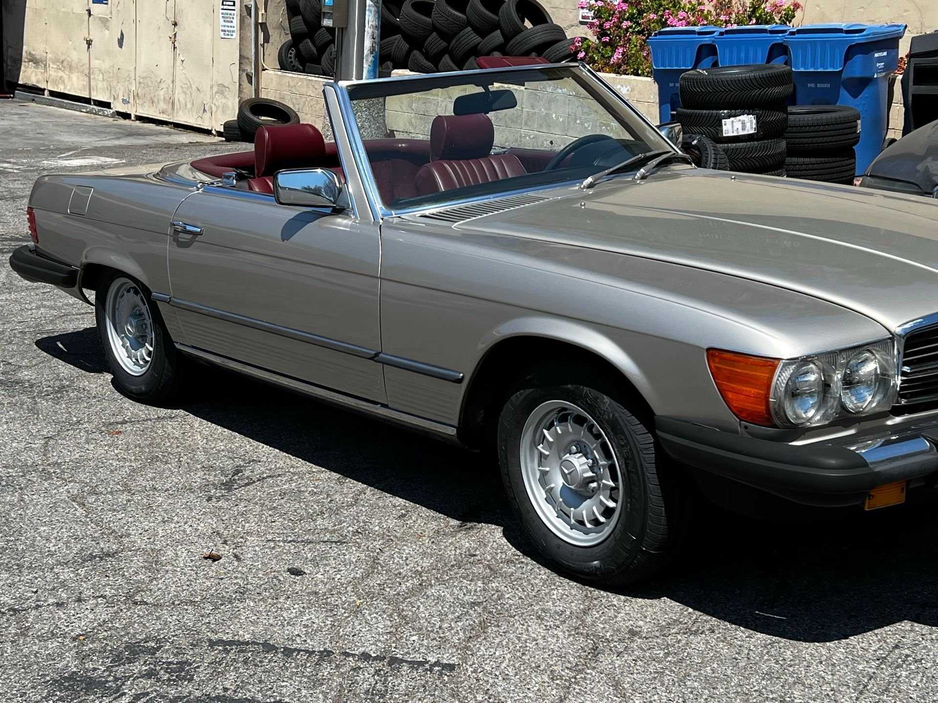A silver Mercedes-Benz R107 convertible parked on an outdoor asphalt lot with maroon seats and classic alloy wheels.