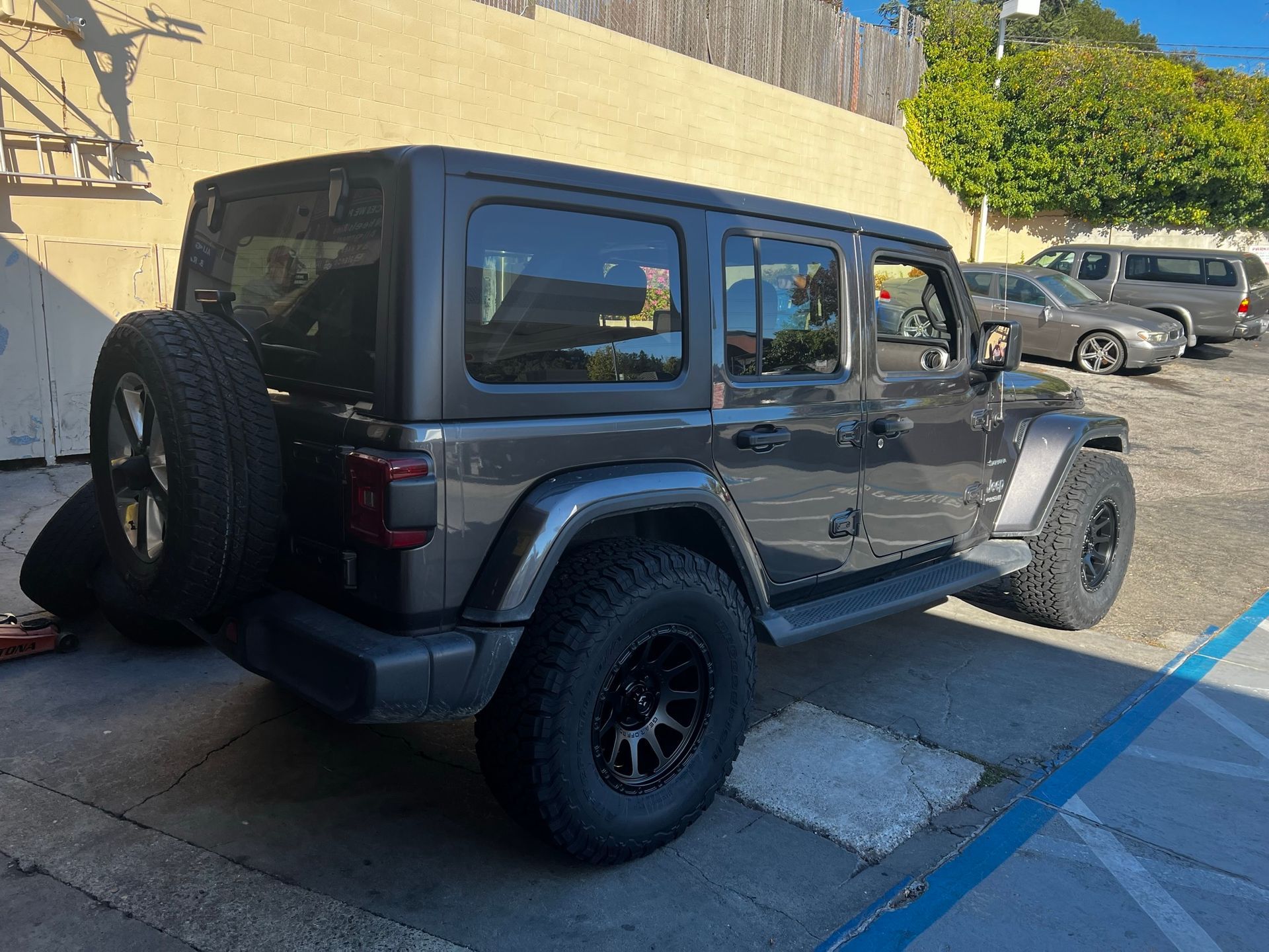 A dark gray Jeep Wrangler parked on asphalt with large off-road tires and a rear-mounted spare wheel.