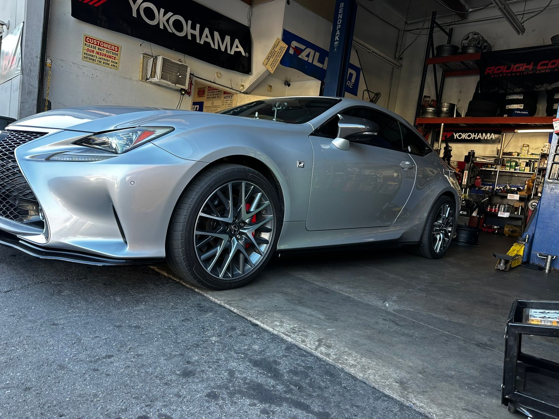 A silver Lexus coupe parked inside an automotive service garage with parts shelves and a Yokohama sign visible.