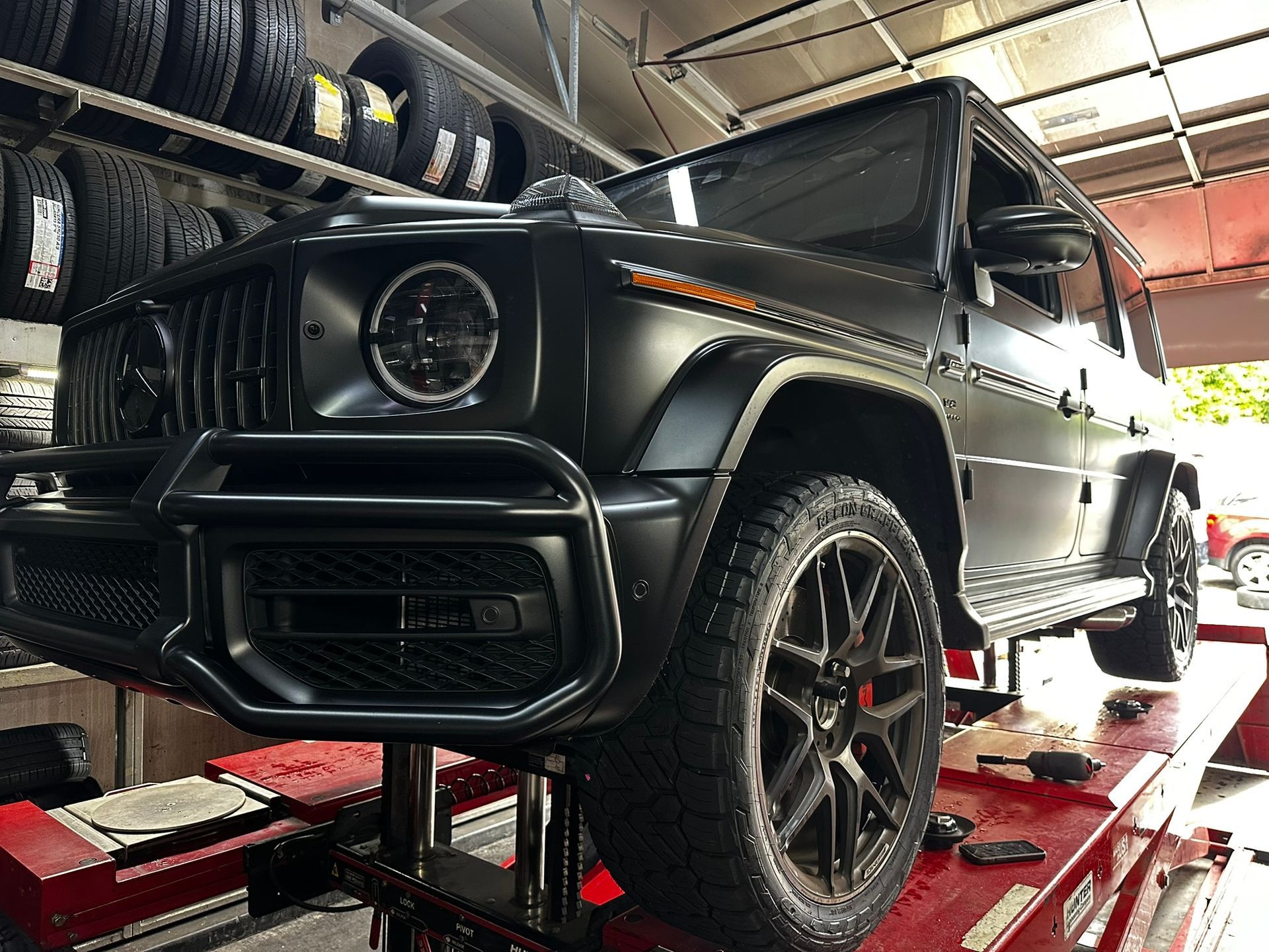 A matte black Mercedes-Benz G-Wagon is elevated on a red automotive lift inside a tire repair shop.