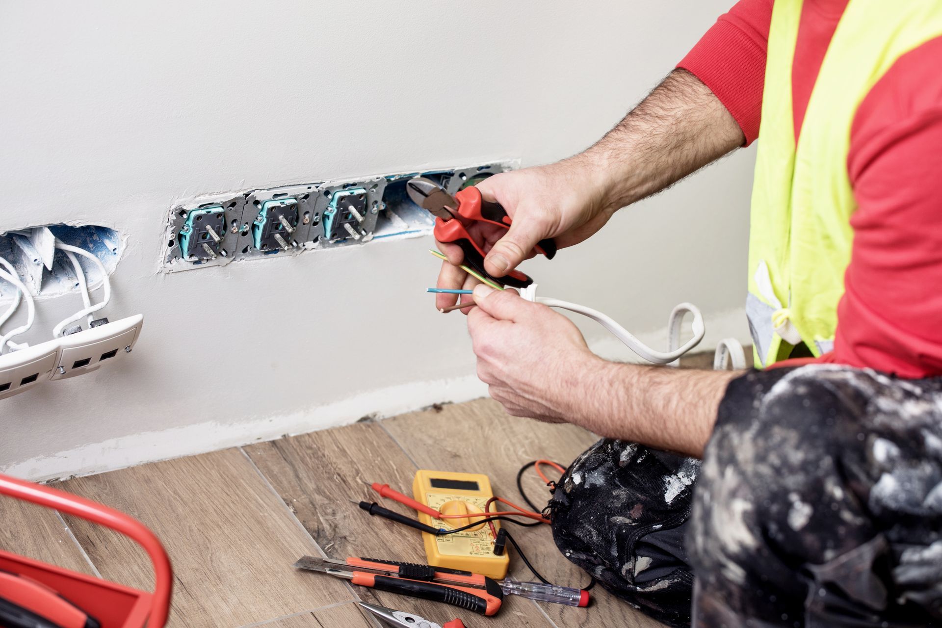 Electrician working on wall outlets, using pliers to strip wire. Yellow vest, tools on floor.