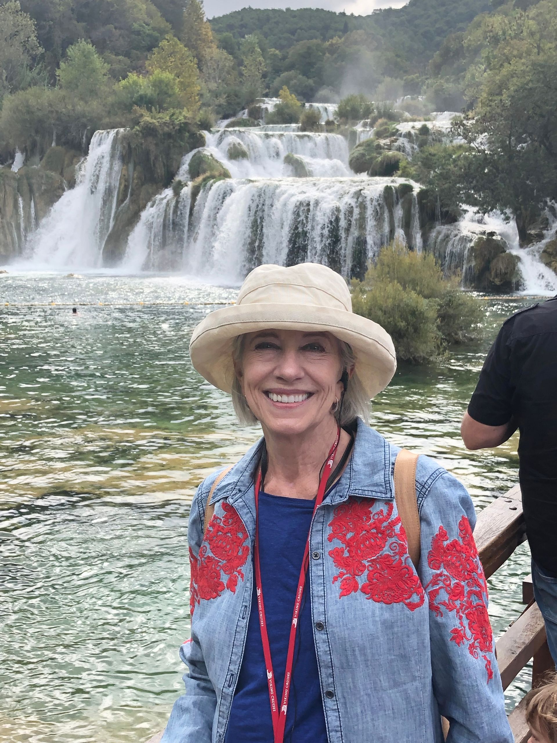A smiling person in a hat and embroidered denim jacket stands in front of the Krka waterfalls in Croatia.