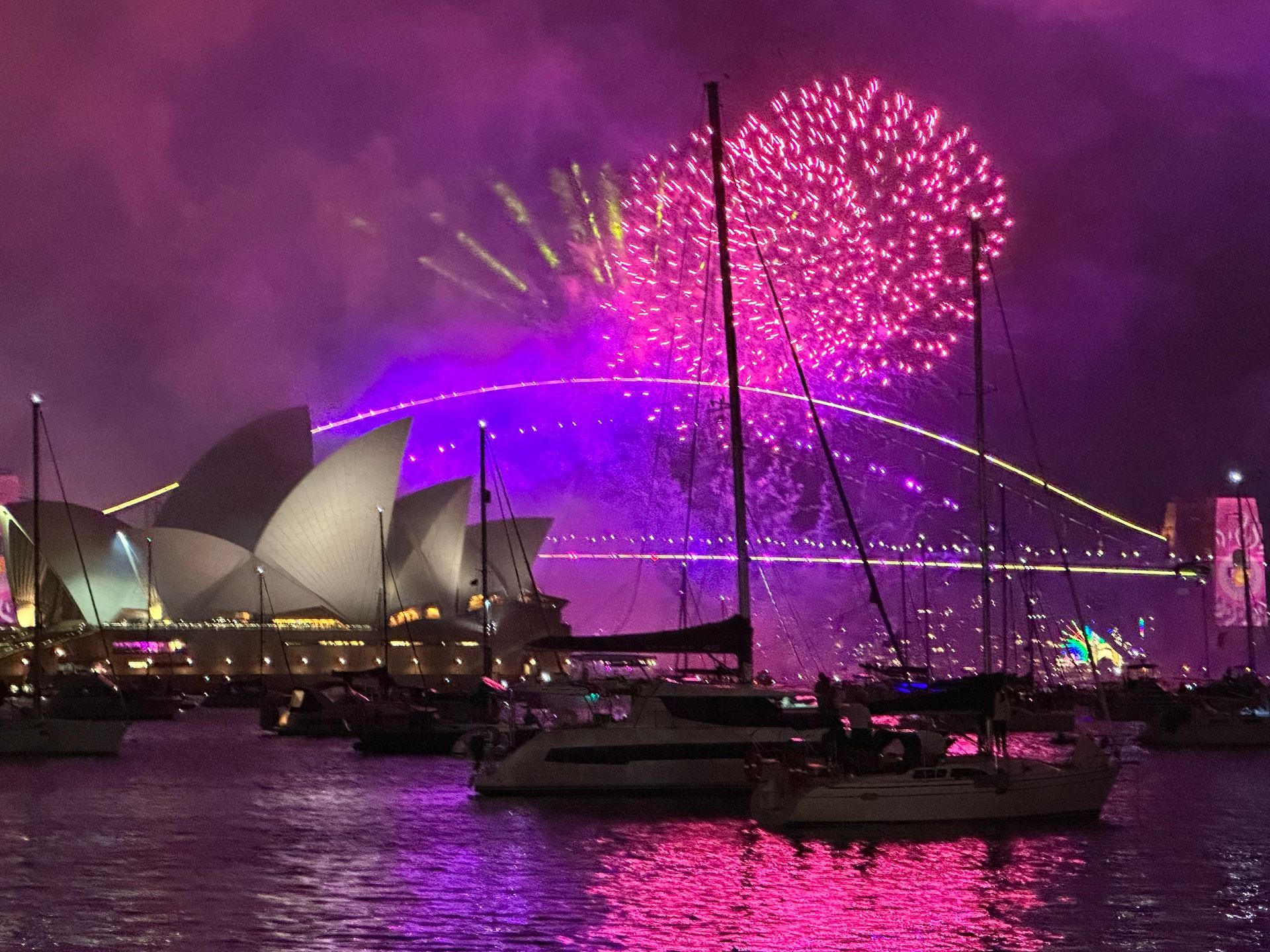 Vibrant purple fireworks explode over the Sydney Opera House and Harbour Bridge, with boats docked in the harbor below on New Year's Eve.