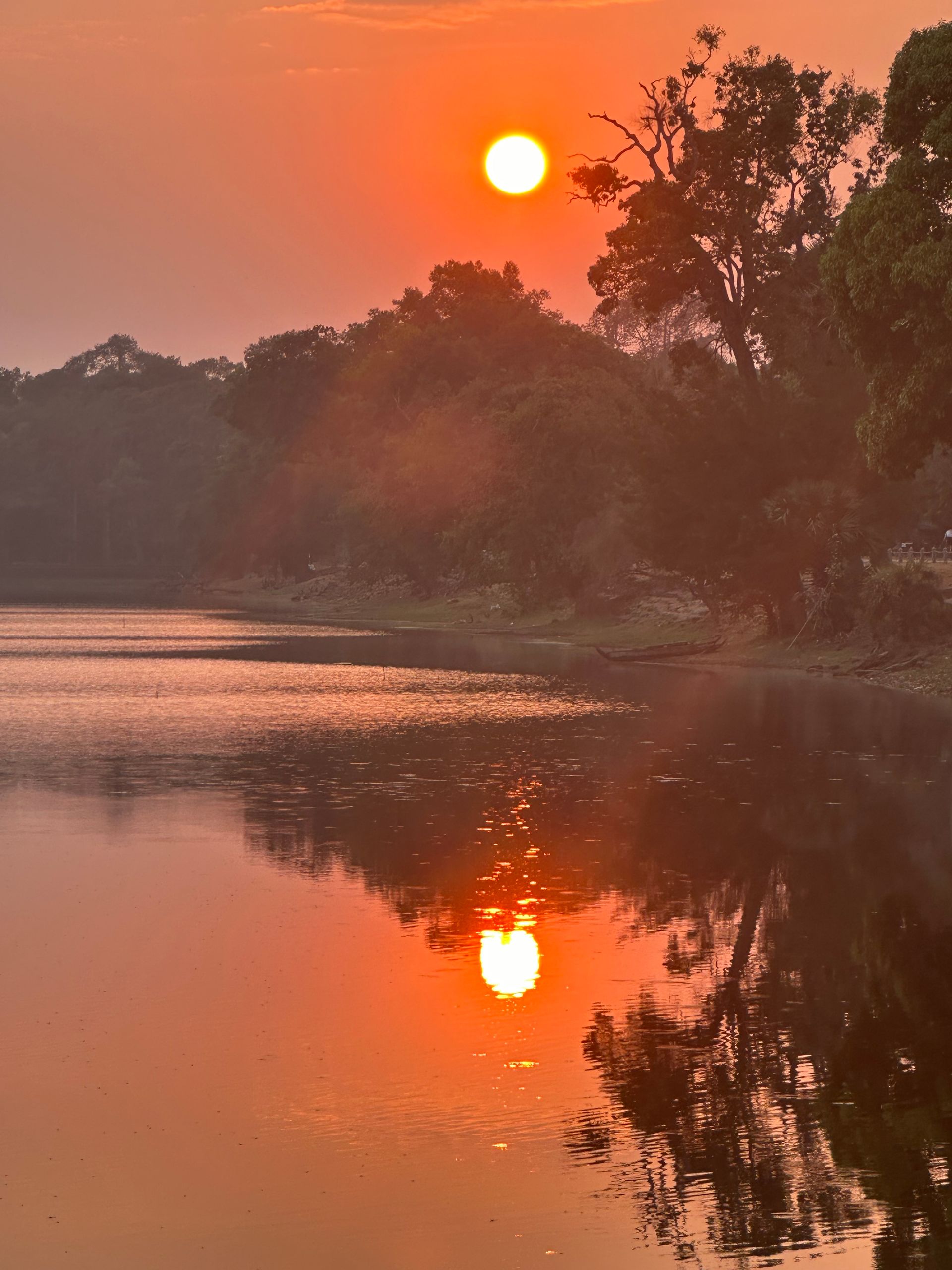 A vibrant orange sunset glows over a calm lake in Siem Reap, casting a bright reflection on the water beside a silhouetted tree line.