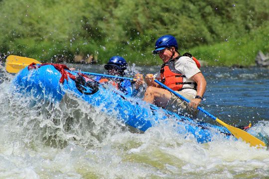 Two people in helmets and life vests paddle a blue raft through splashing river rapids.