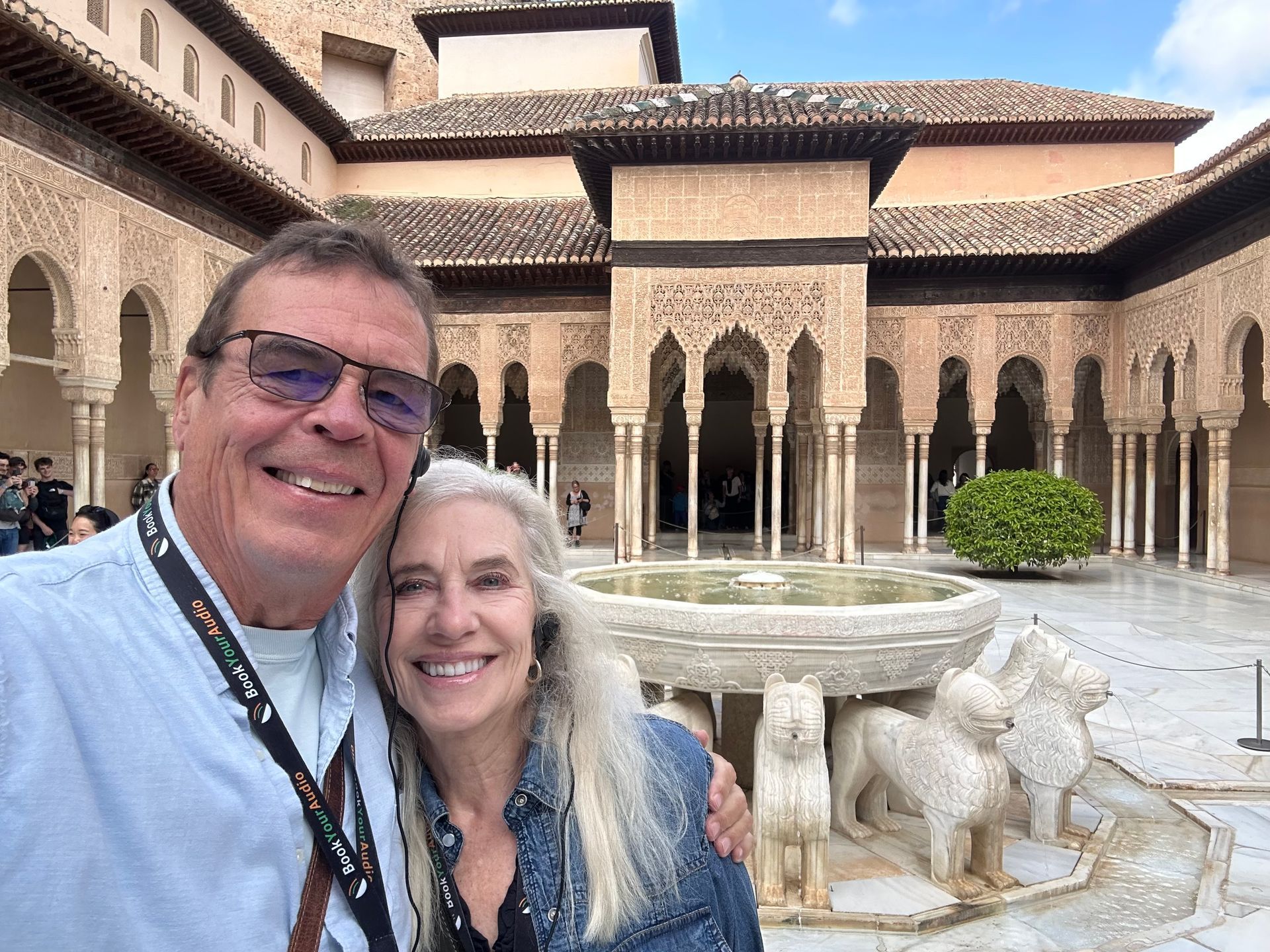 A smiling couple poses for a selfie in front of the ornate Lion Fountain in the courtyard of the Alhambra, Spain.