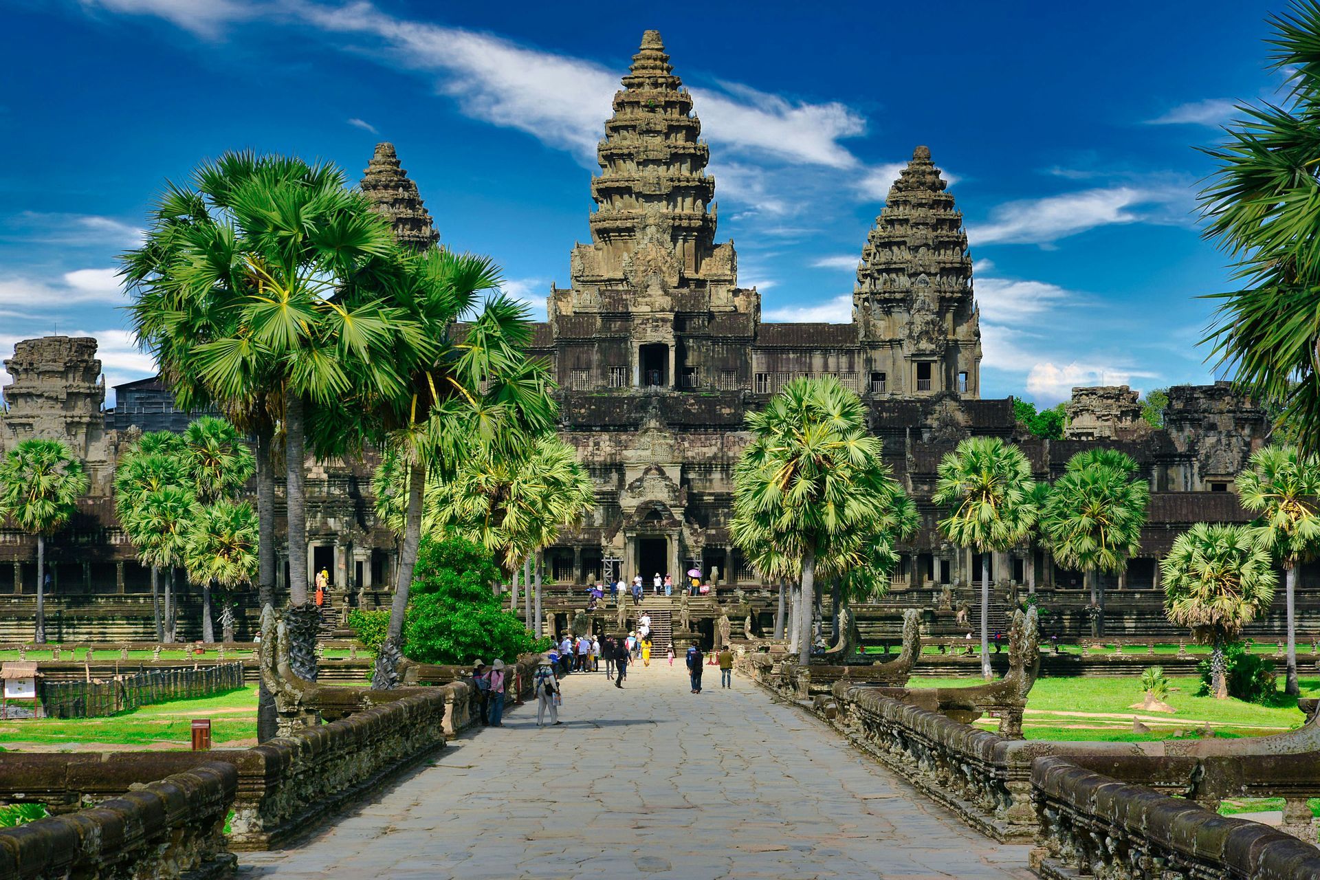 Angkor Wat temple entrance with stone towers, palm trees, and a walkway under a bright blue sky