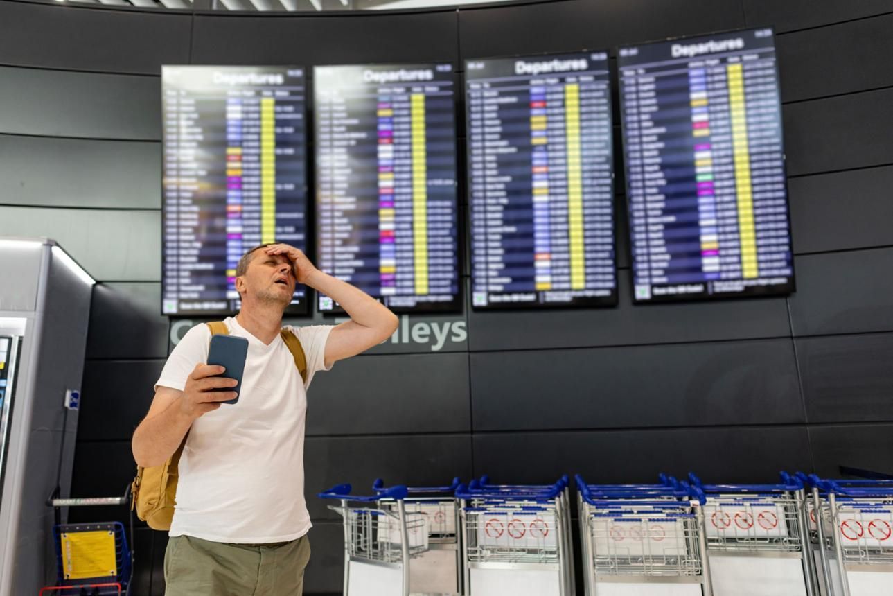 A person is standing in front of an airport flight board, holding a cellphone and 