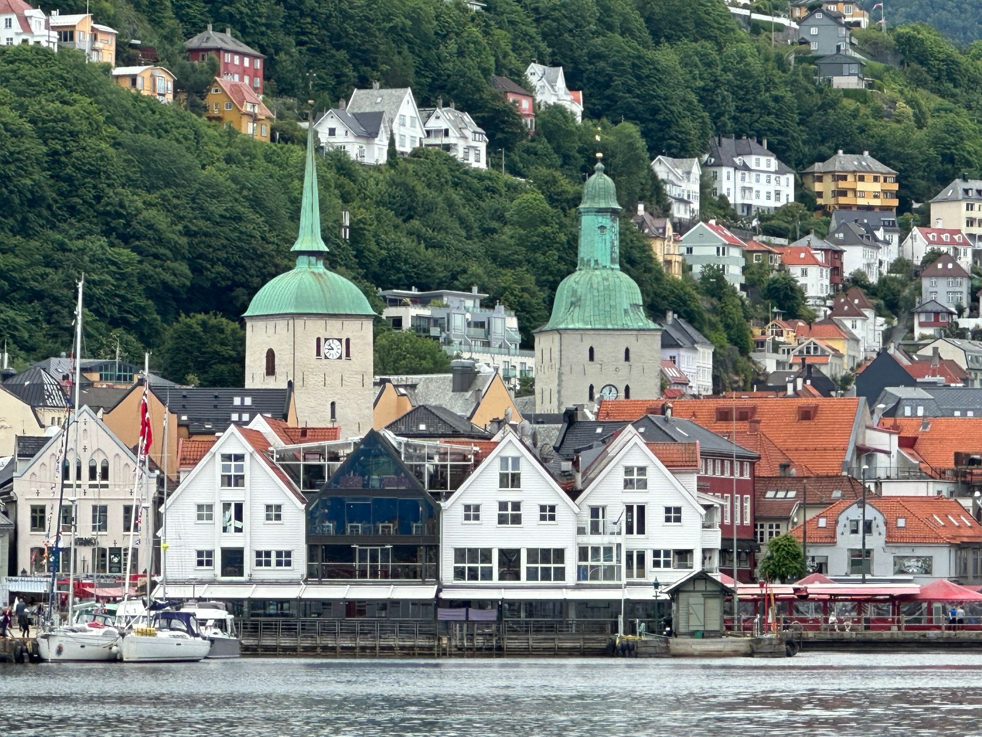 Bergen, Norway: A waterfront view of Bryggen’s white historic wooden buildings, two church towers, and hillside houses.