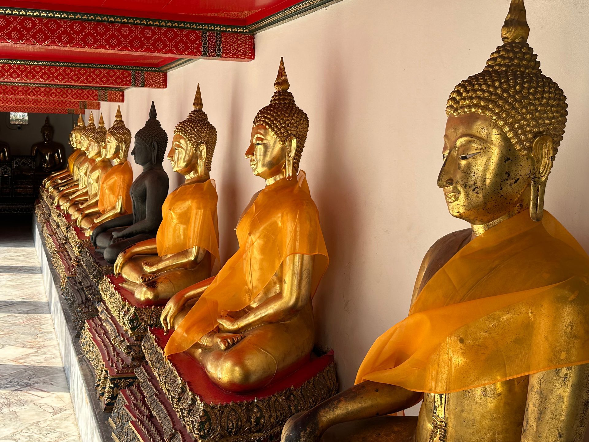 A row of golden Buddha statues wearing yellow robes, seated in a sheltered, red-roofed corridor.
