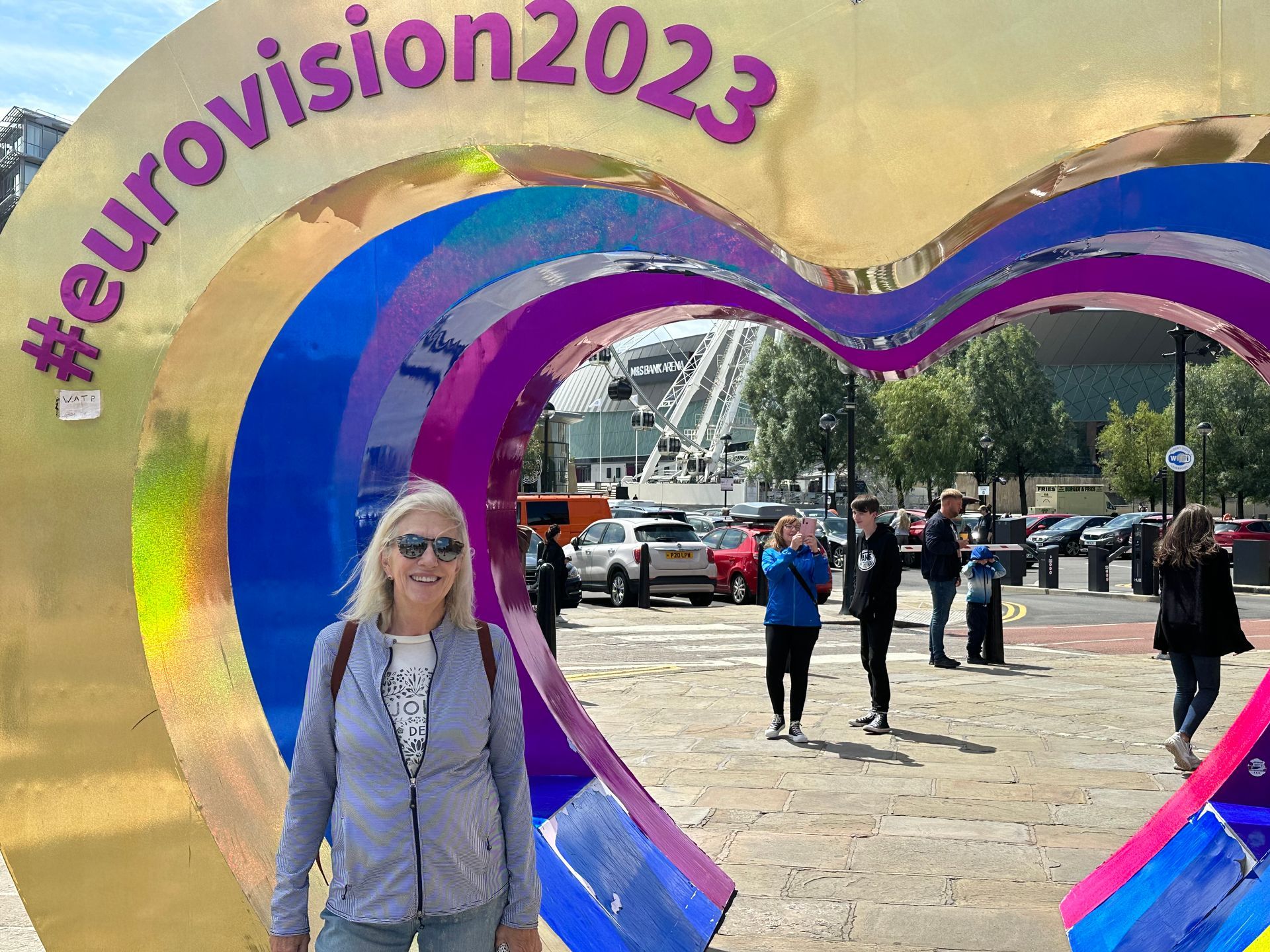 A smiling woman stands in front of a large, heart-shaped Eurovision 2023 sign in a sunny outdoor plaza.