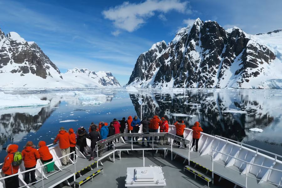 People in red parkas are on the bow of an expedition cuise ship in Antarctica with snow-covered mountains in the background.