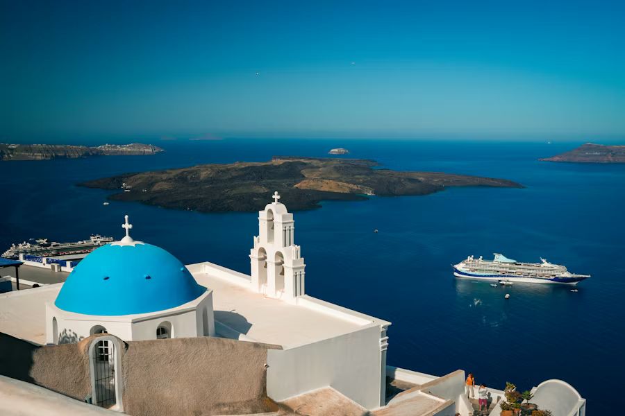 Blue-domed church overlooking a seaside bay with a yacht and islands under a clear sky.