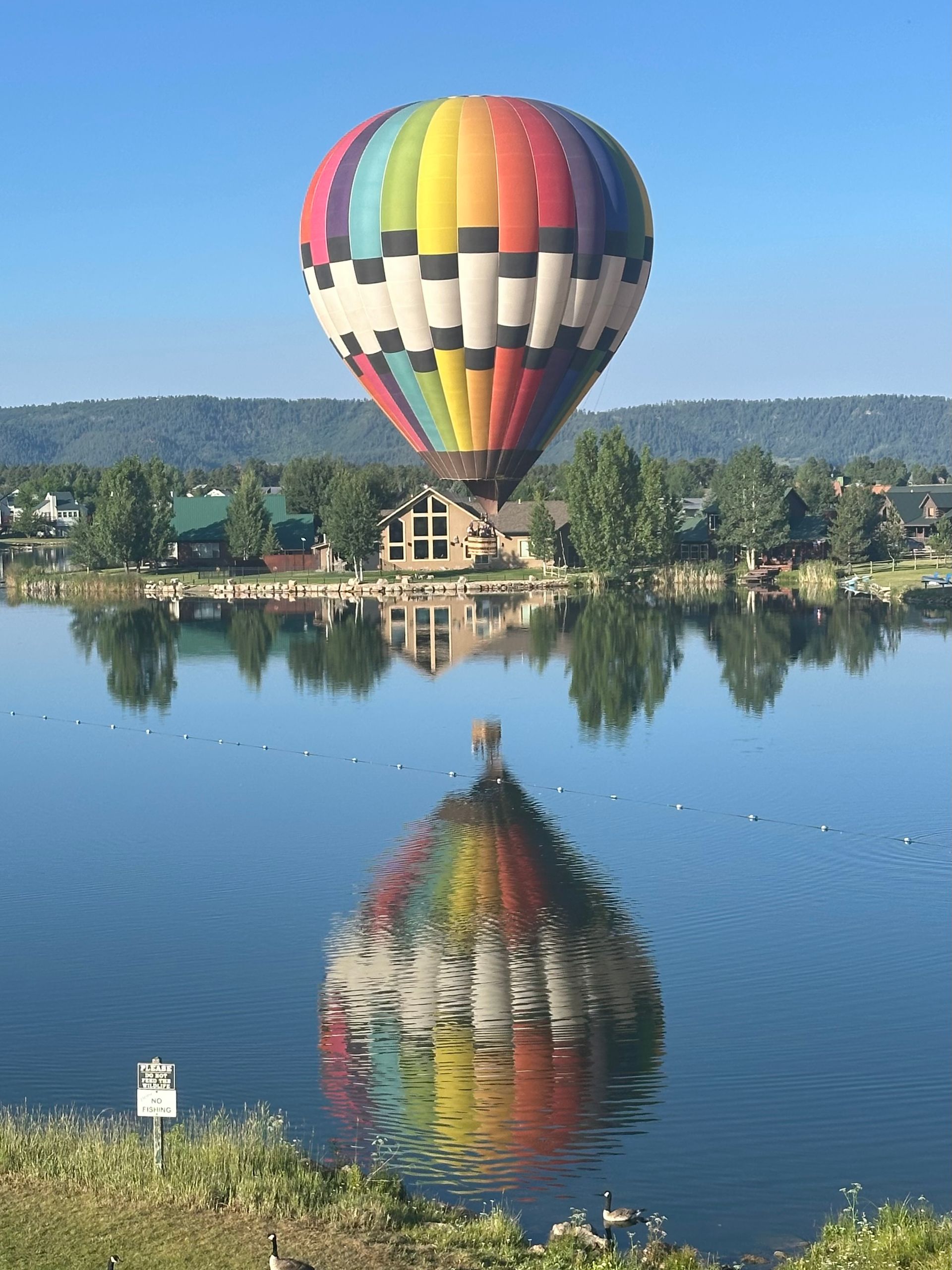 A colorful hot air balloon floats over a calm lake, creating a vibrant reflection on the water's surface.
