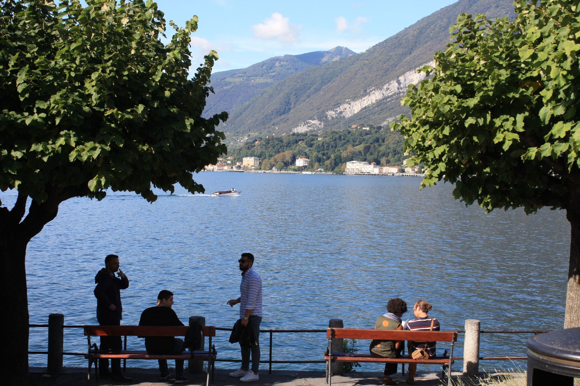 People relax on benches by Lake Como, framed by two large trees with a mountain and buildings in the background.