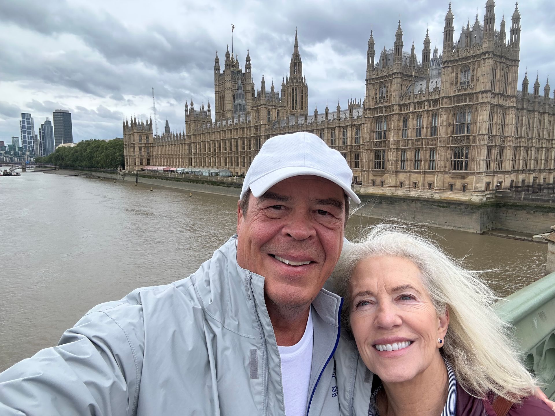 A smiling couple takes a selfie on a bridge in London, with the Palace of Westminster in the background.