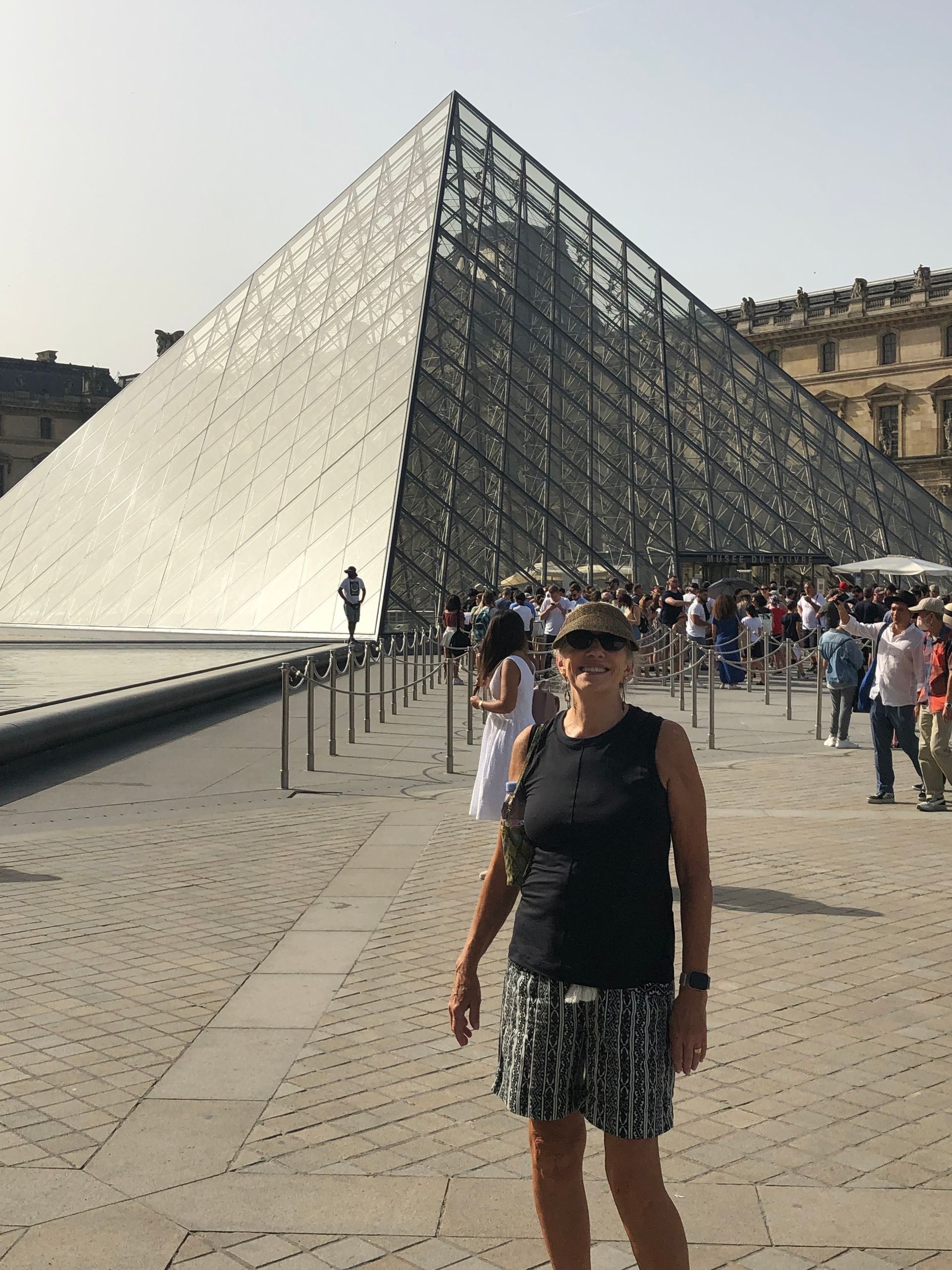 A person in a hat and dark top stands smiling in front of the glass pyramid at the Louvre Museum in Paris.