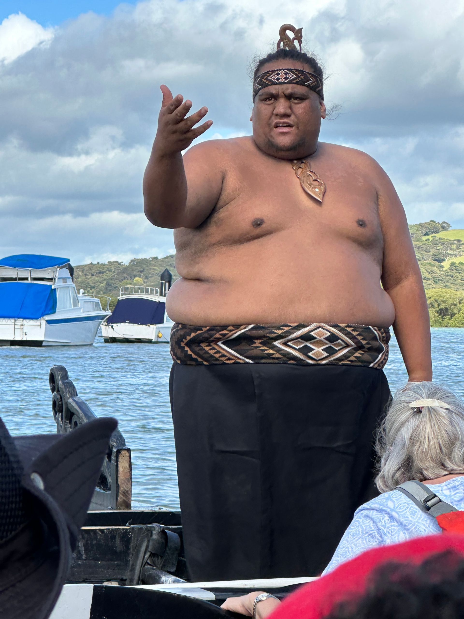 A New Zealand  wearing a patterned headband and sash stands on a boat on the water, gesturing with their hand while speaking.