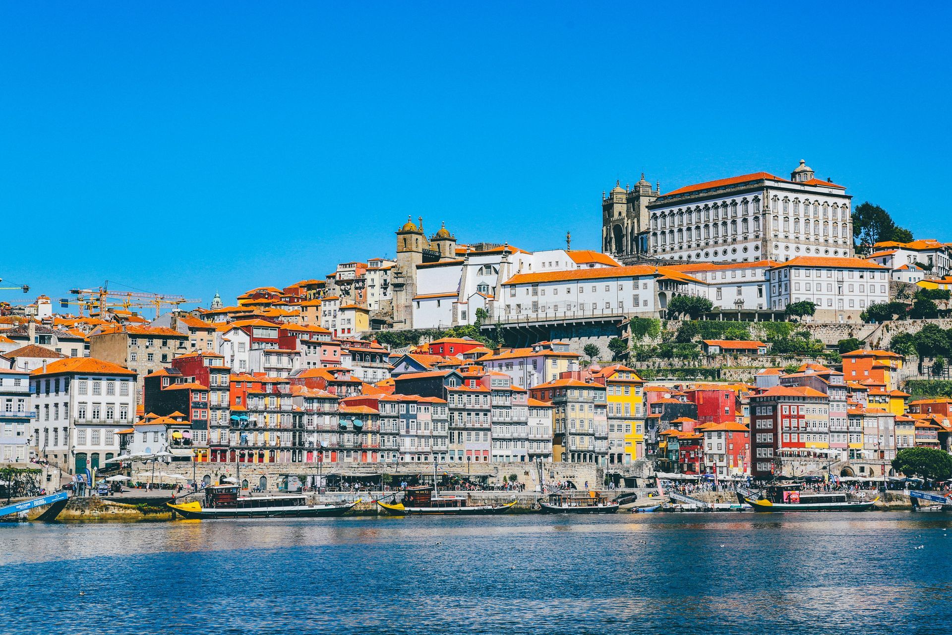 Colorful hillside cityscape with red-roofed buildings overlooking a blue waterfront under a clear sky