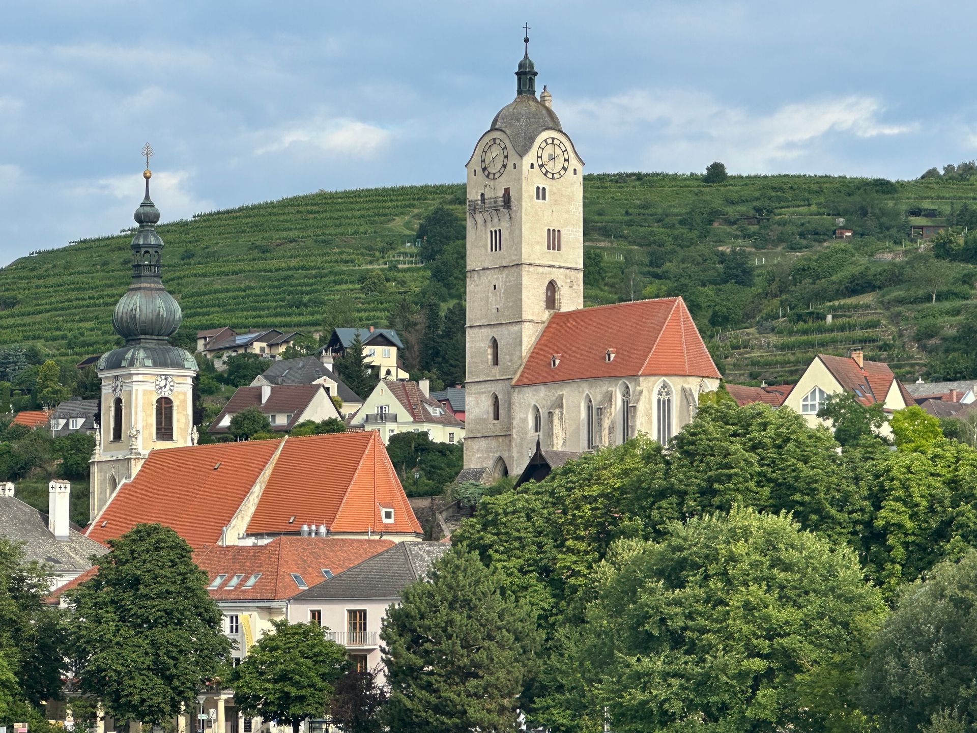 Two stone churches with red roofs sit nestled at the base of a green, terraced hillside under a blue, cloudy sky.