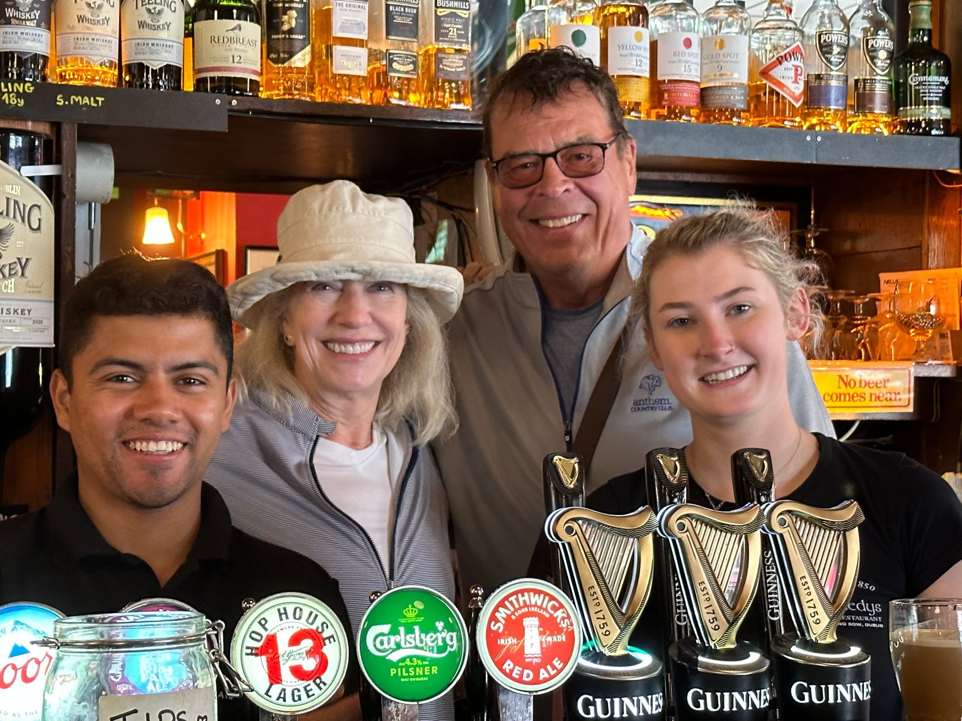 Four people smiling behind a bar featuring beer taps and a back bar shelf filled with various bottles of liquor.