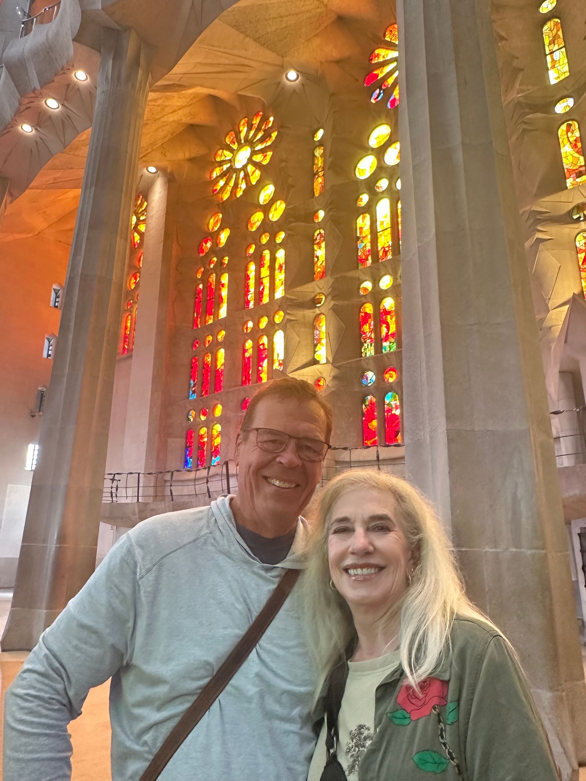 A smiling couple stands inside the Sagrada Família, framed by massive stone columns and vibrant stained-glass windows.