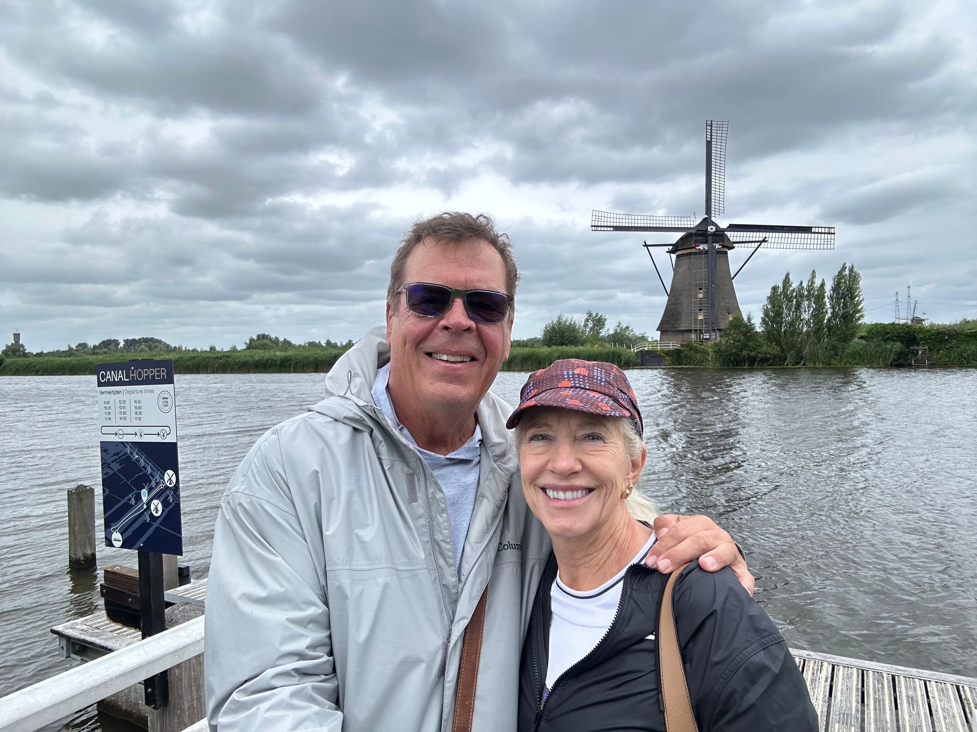 A smiling couple stands on a pier with a windmill and a cloudy sky in the background.