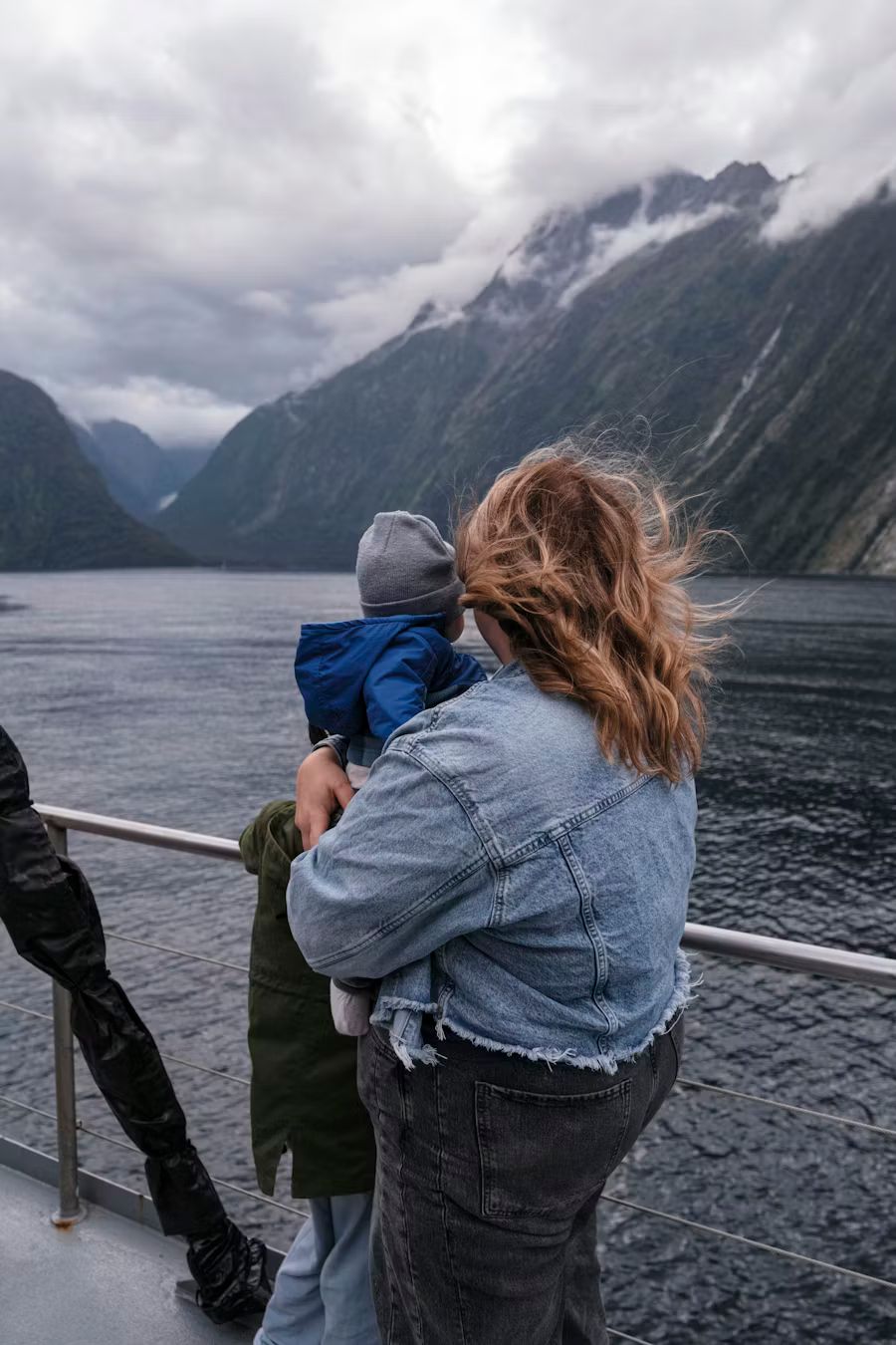 A mother and child have their backs to the camera, looking out over the water and mountains from a cruise ship