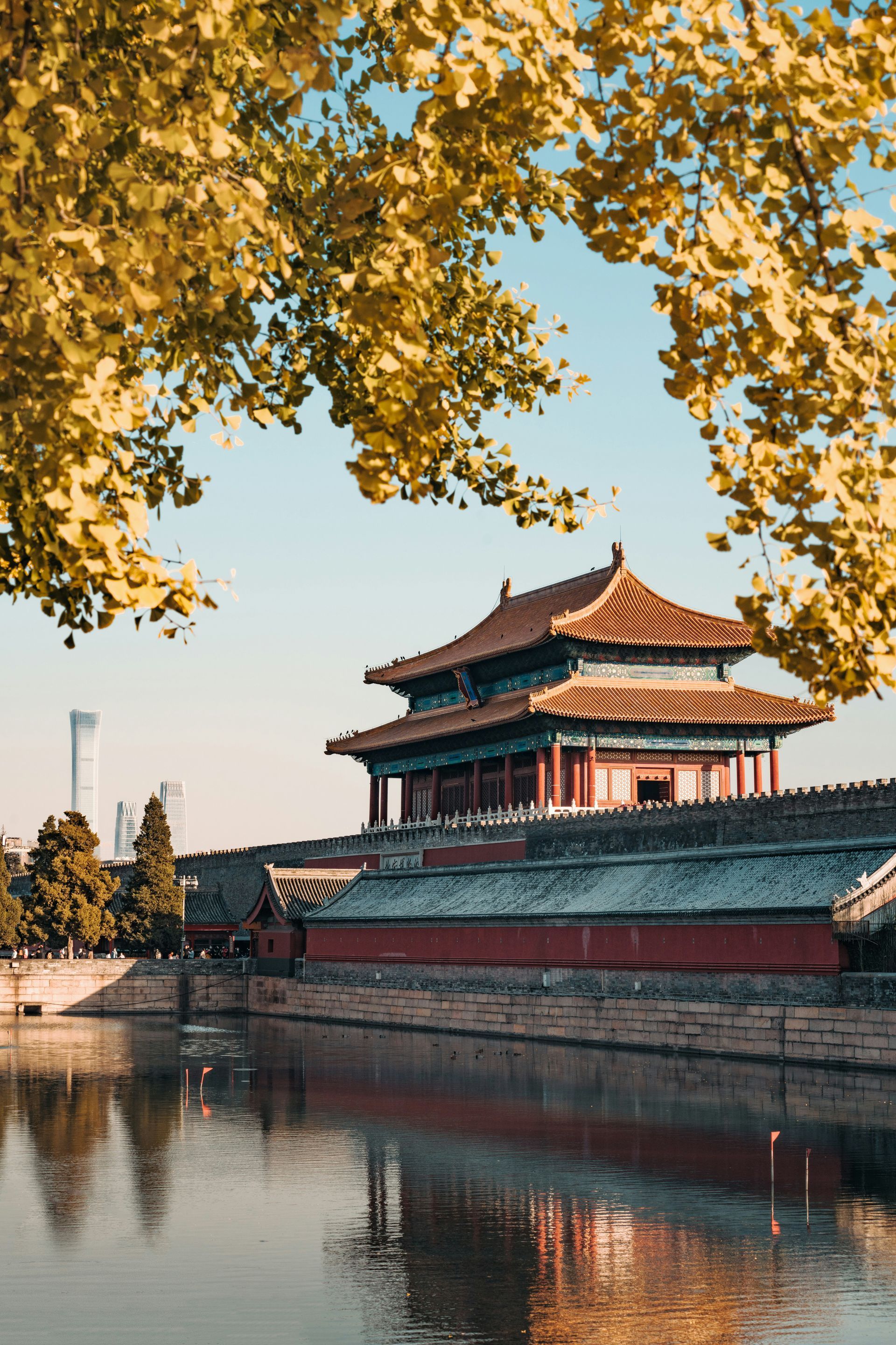 Historic Chinese palace by a moat, framed by golden autumn leaves under a clear blue sky