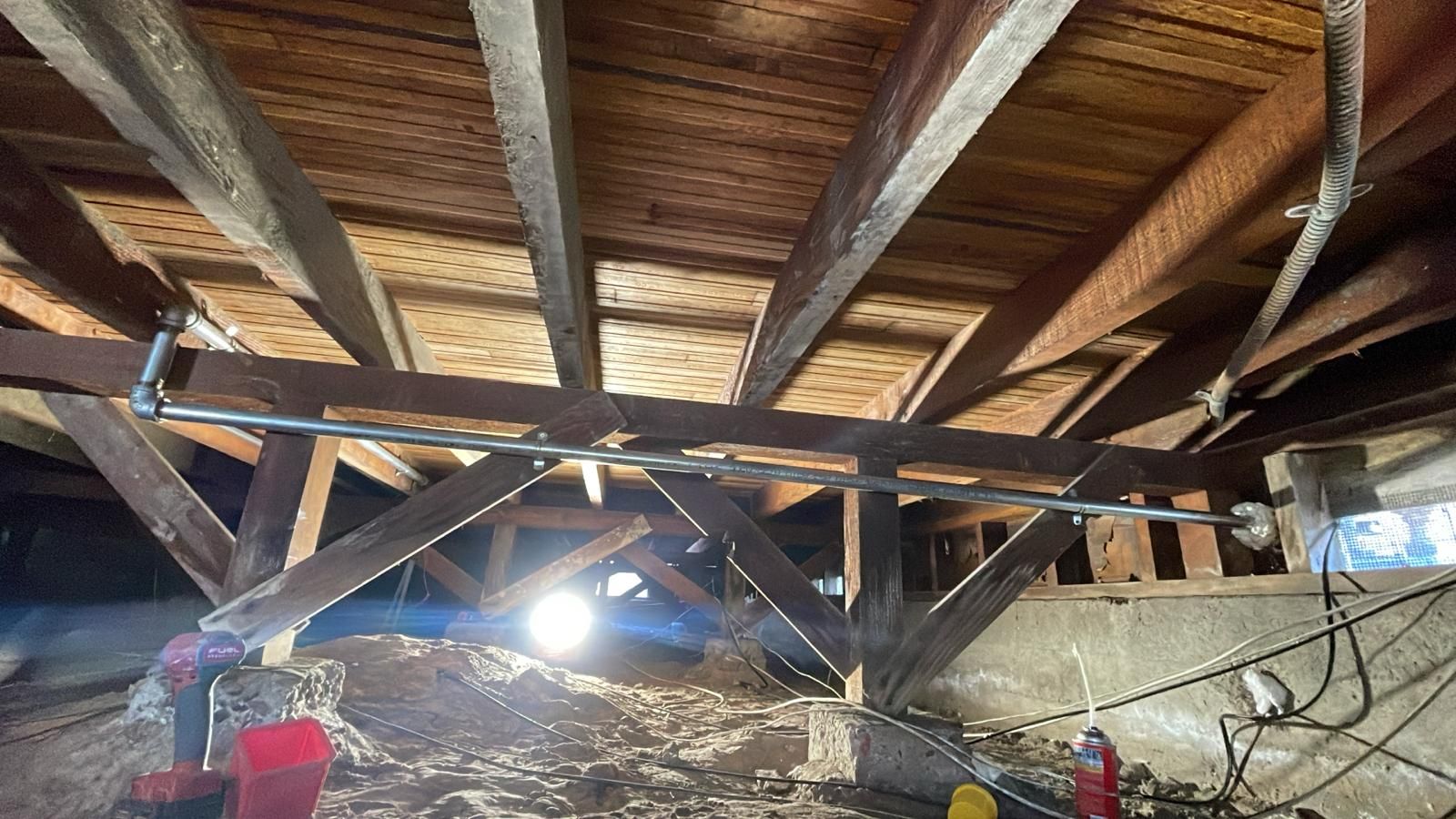 A metal pipe support is installed beneath wooden floor joists in a dimly lit crawlspace.
