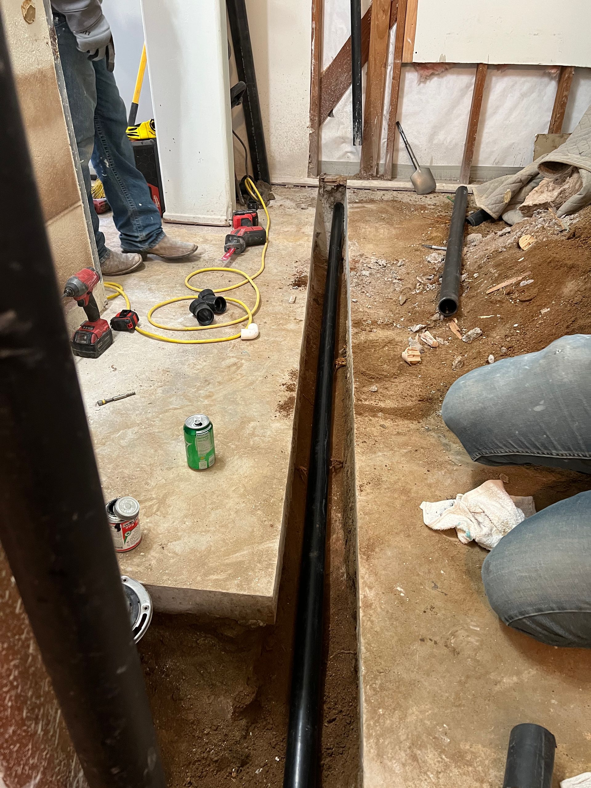 Construction workers installing a black drainage pipe in a trench cut through a concrete floor.