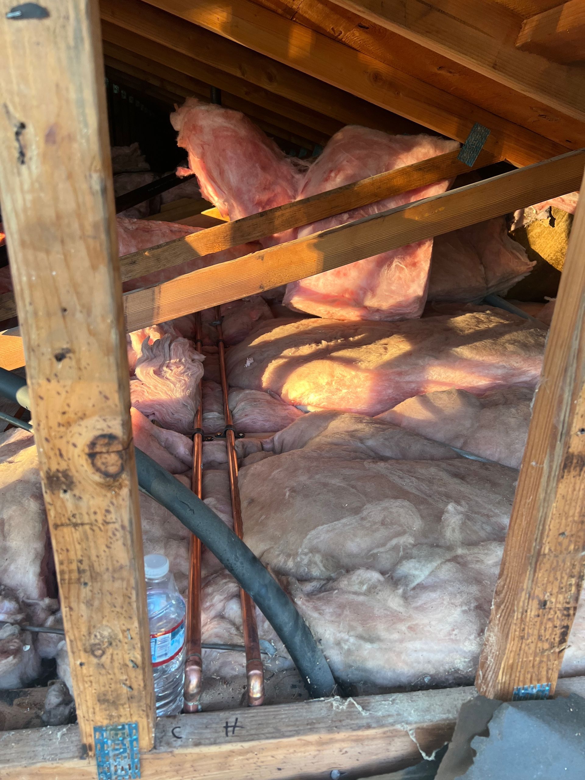 Copper pipes run through fiberglass insulation inside an attic, framed by wooden roof rafters.
