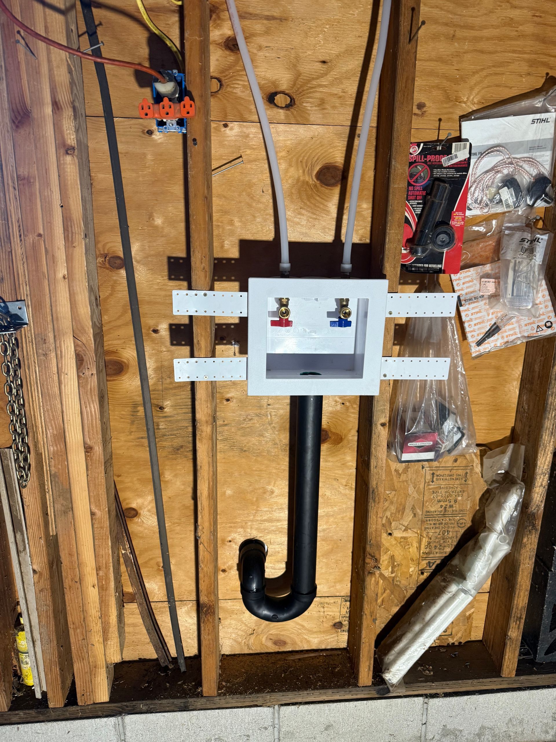 Washing machine hookup box with supply lines and a black drain pipe mounted between wooden wall studs.