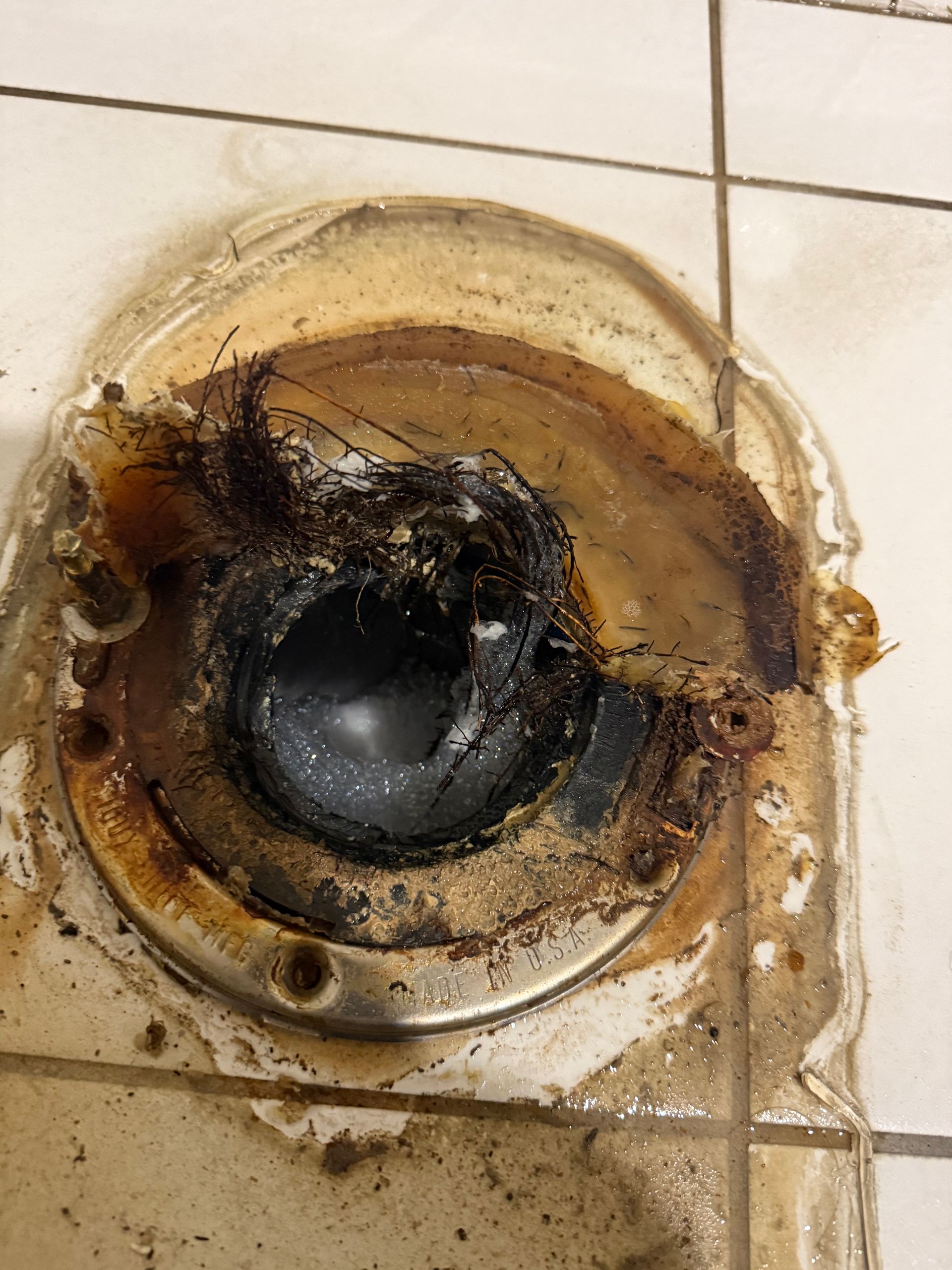 A toilet flange on a tiled bathroom floor, showing signs of buildup, rust, and debris around the drain opening.