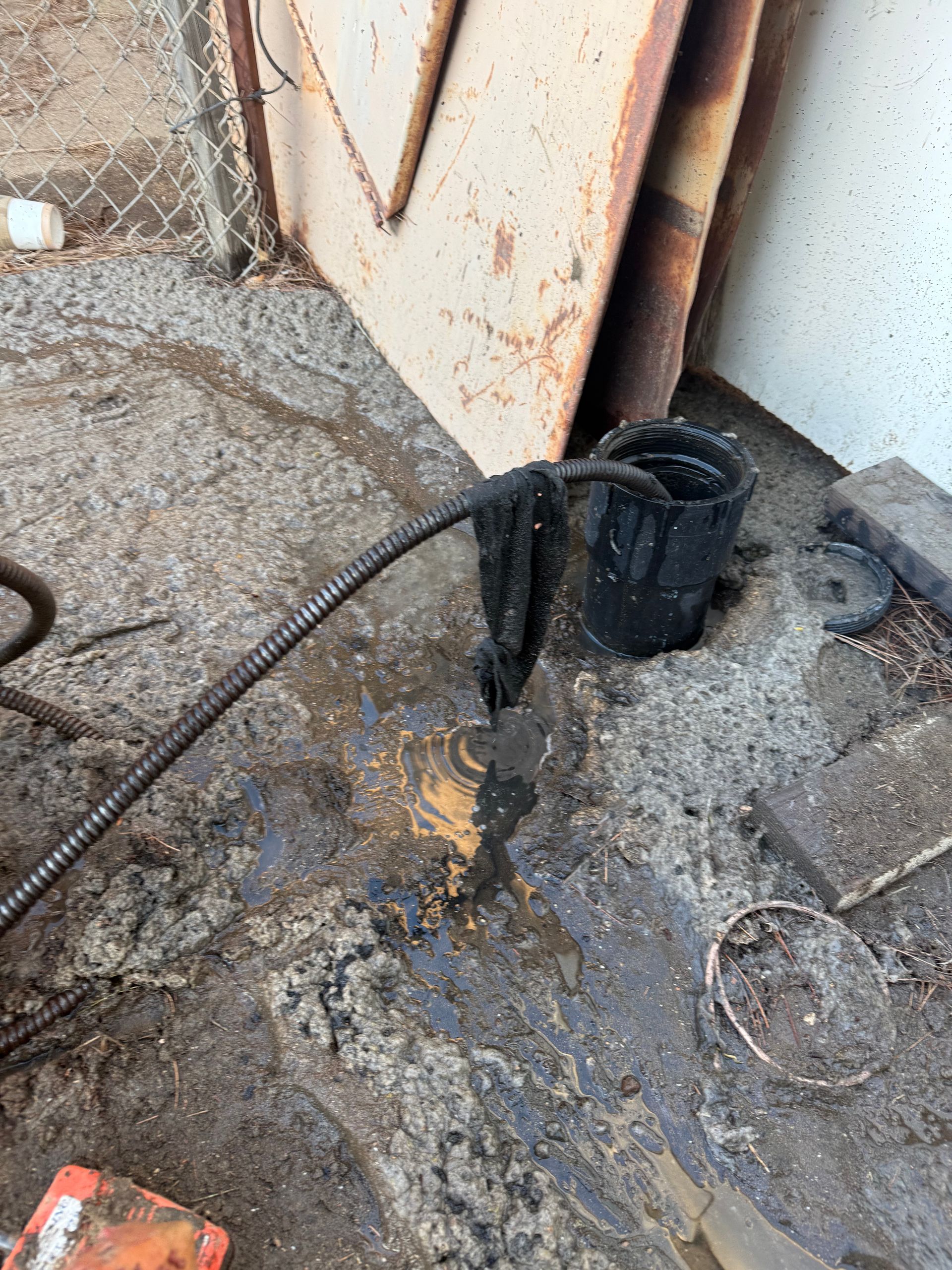 A black drainage hose snakes from a pipe into a bucket, leaking dark sludge onto the surrounding dirt and concrete.