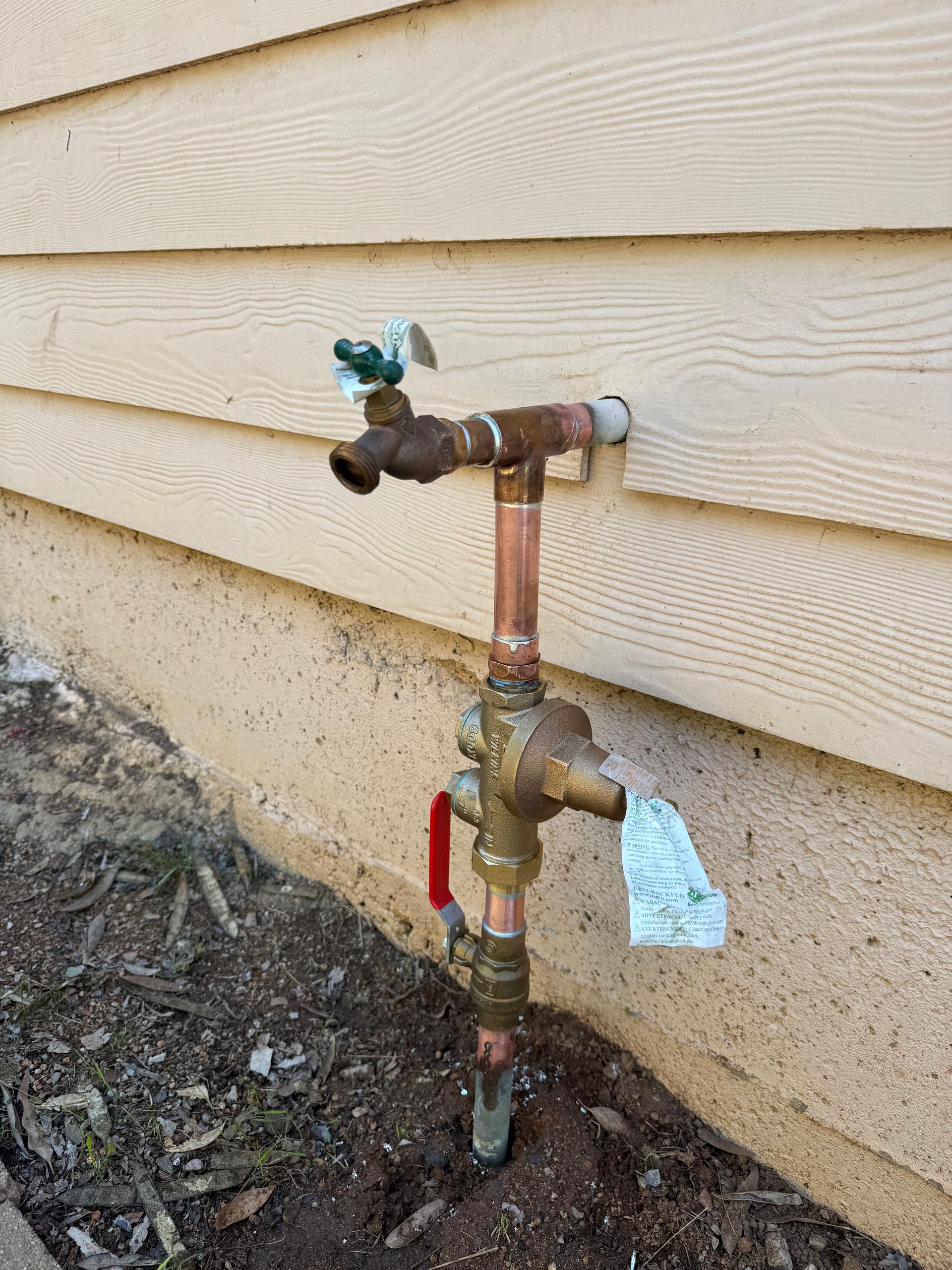 An exterior copper plumbing assembly with a water spigot and a red-handled shutoff valve mounted against house siding.