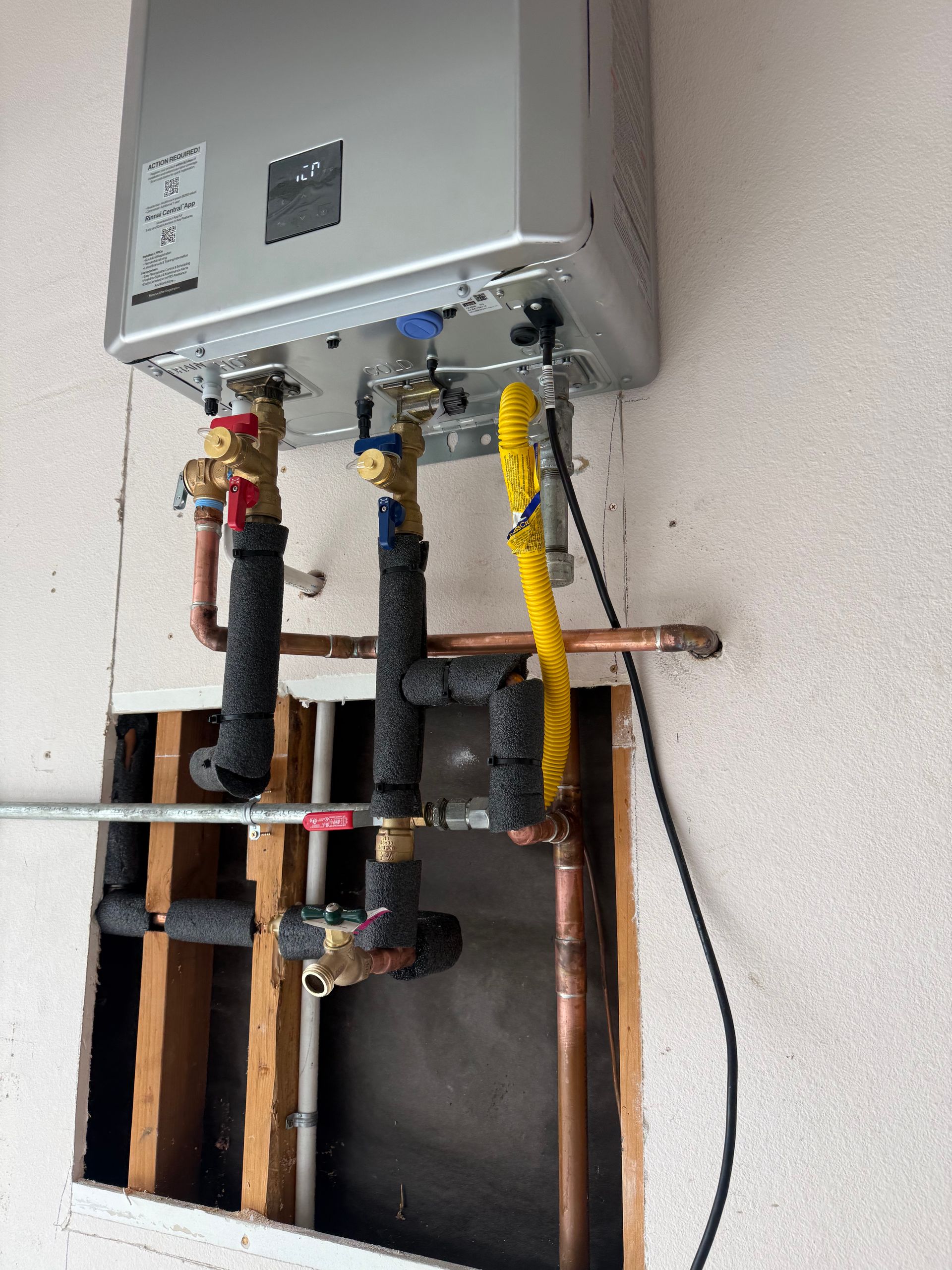 A tankless water heater mounted on a wall with exposed copper pipes, insulated plumbing, and a yellow gas line connection.