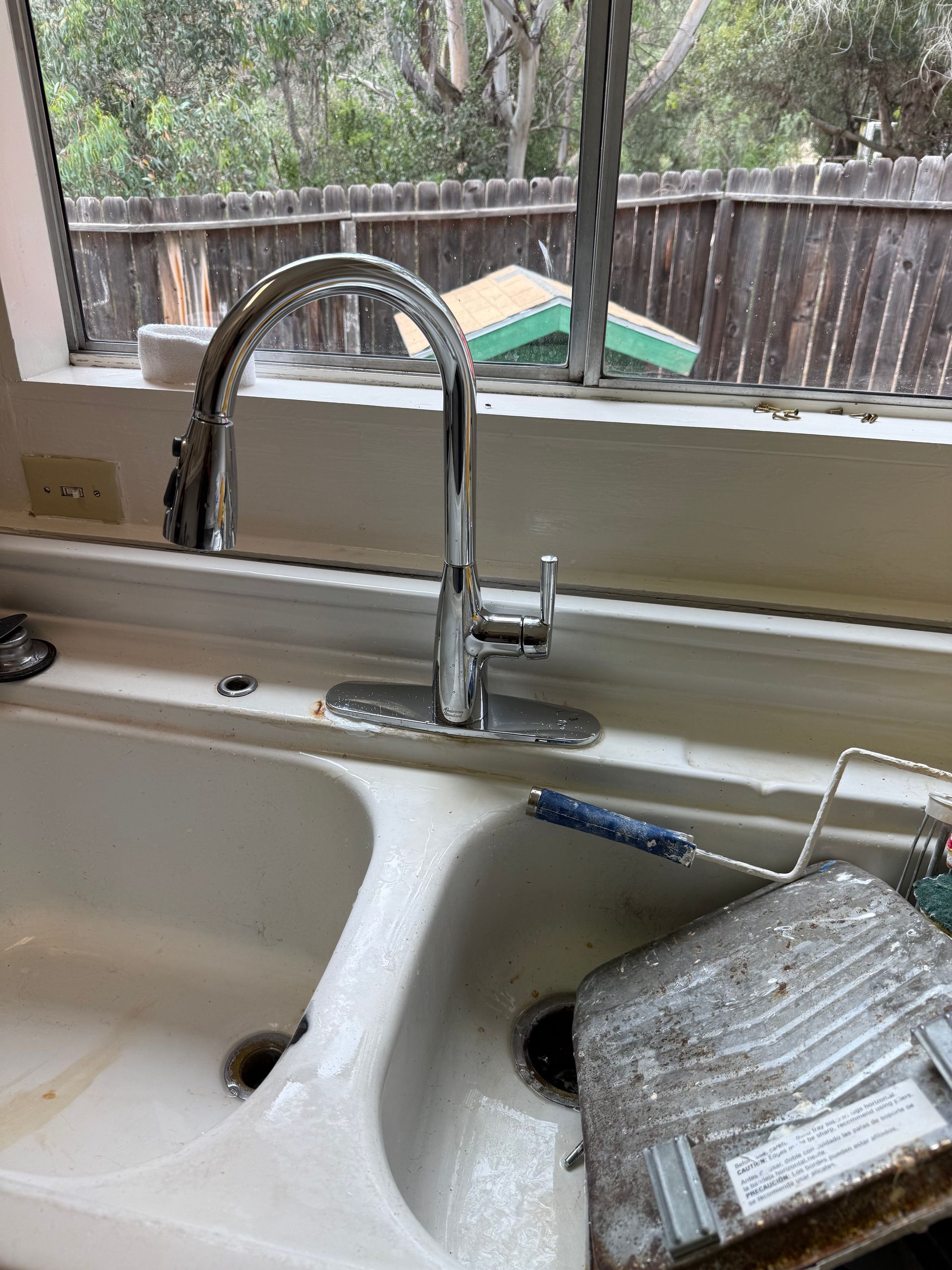 A modern chrome kitchen faucet sits above a white double-basin sink containing a used paint roller tray.