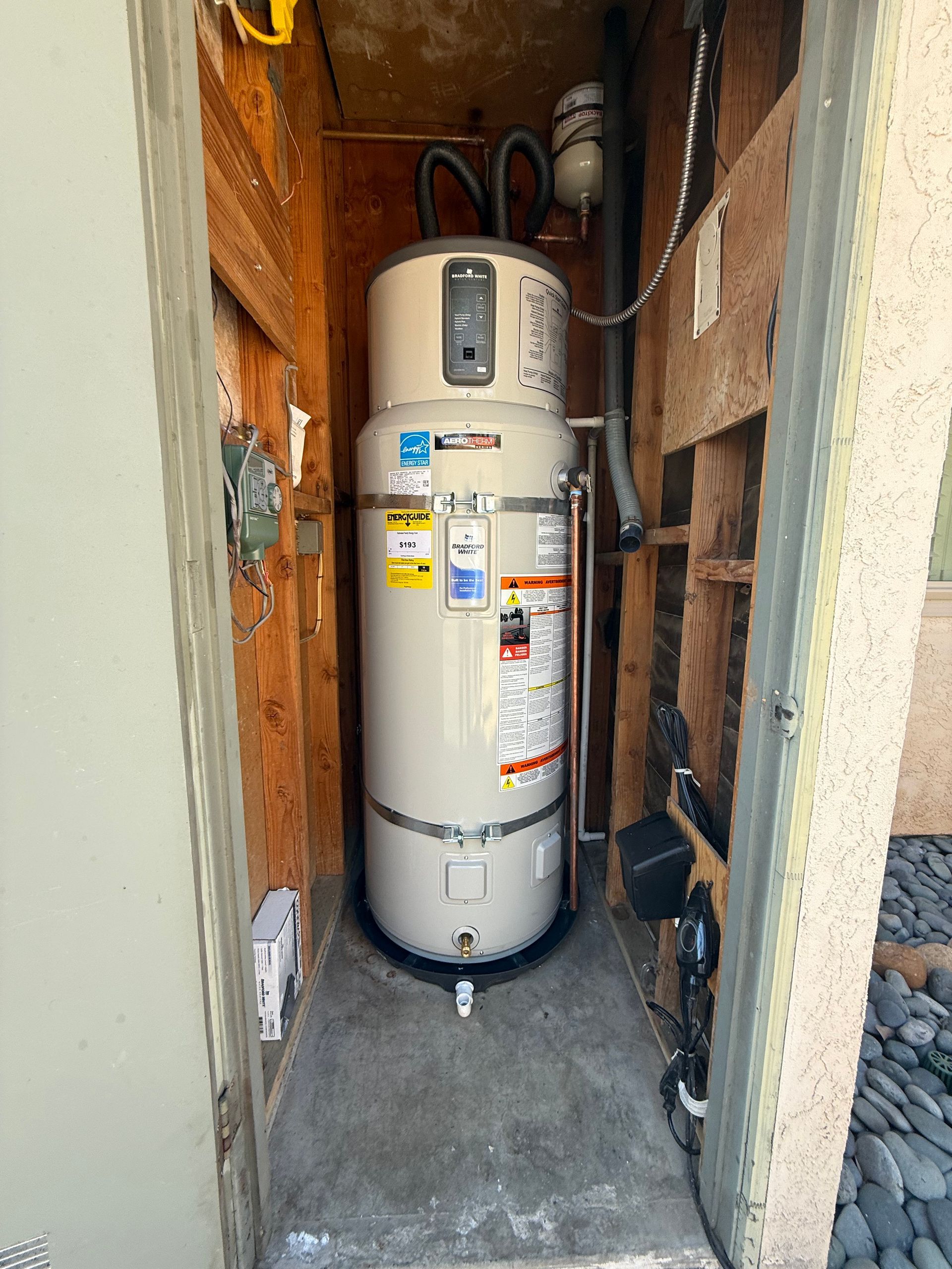 A tall, cream-colored electric water heater stands inside an open, narrow outdoor utility closet with wooden walls.