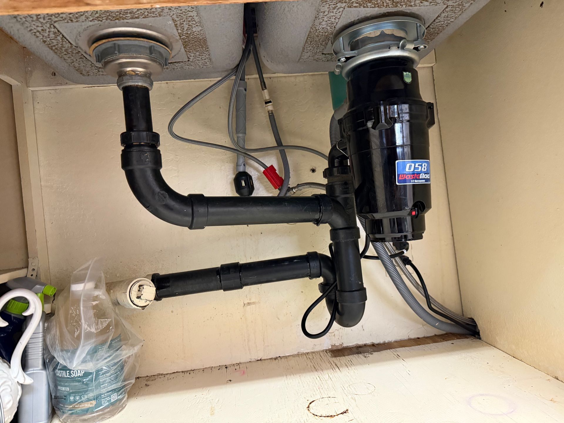 A black sink disposal unit and PVC piping connected to a dual-basin kitchen sink inside a white cabinet.