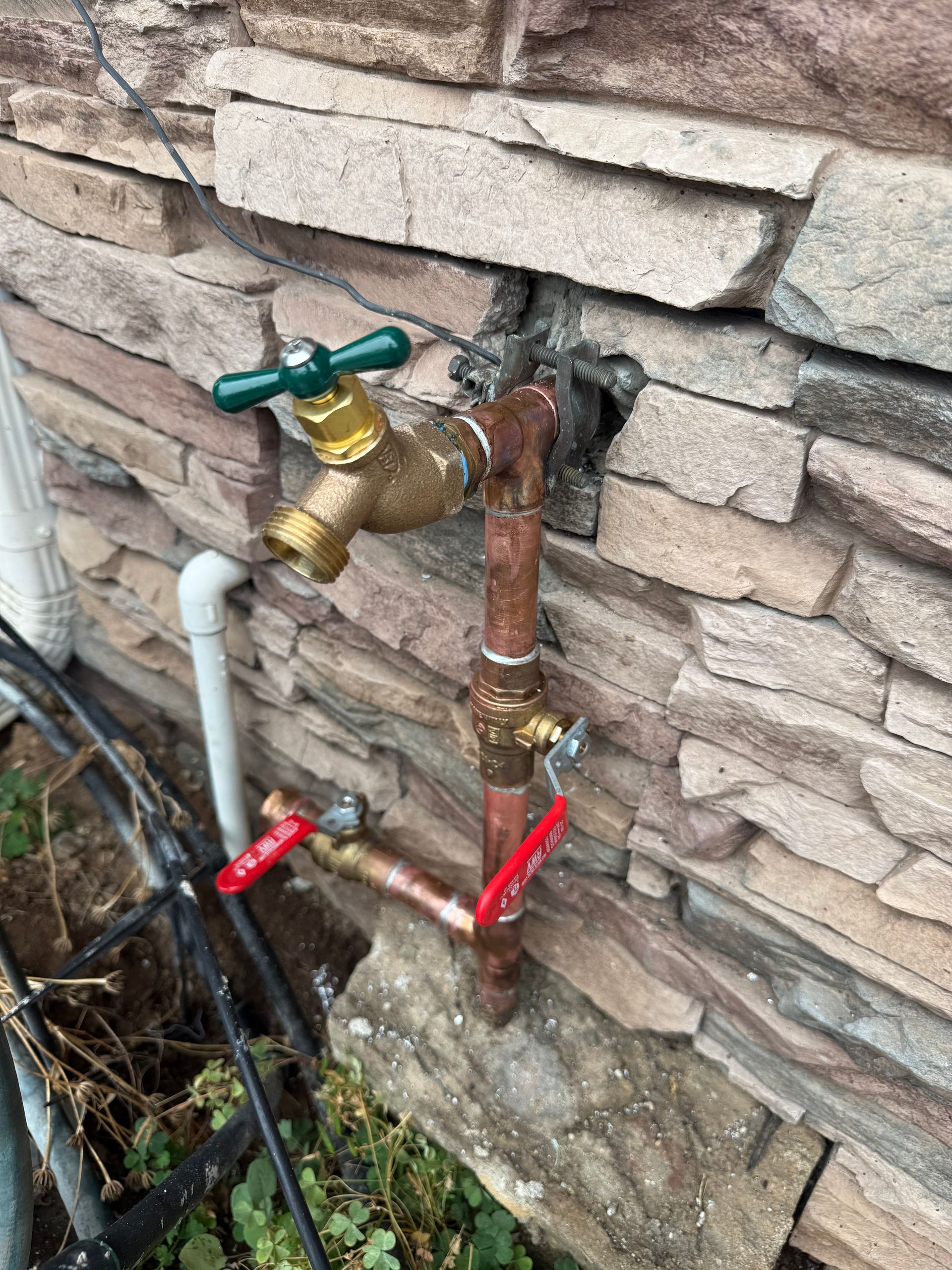 An outdoor copper pipe assembly against a stone wall featuring a green-handled faucet and two red-handled valves.