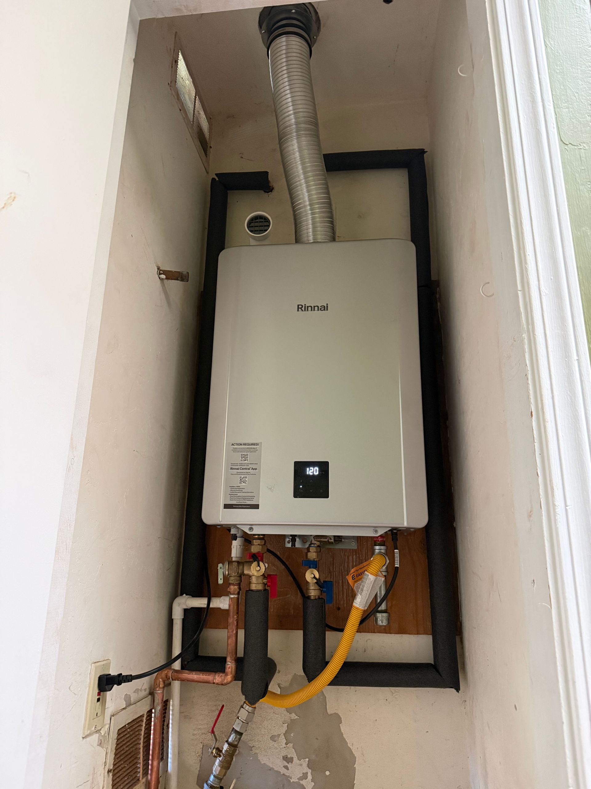 A wall-mounted tankless water heater in a narrow utility alcove with exposed pipes, insulation, and a vent pipe above.