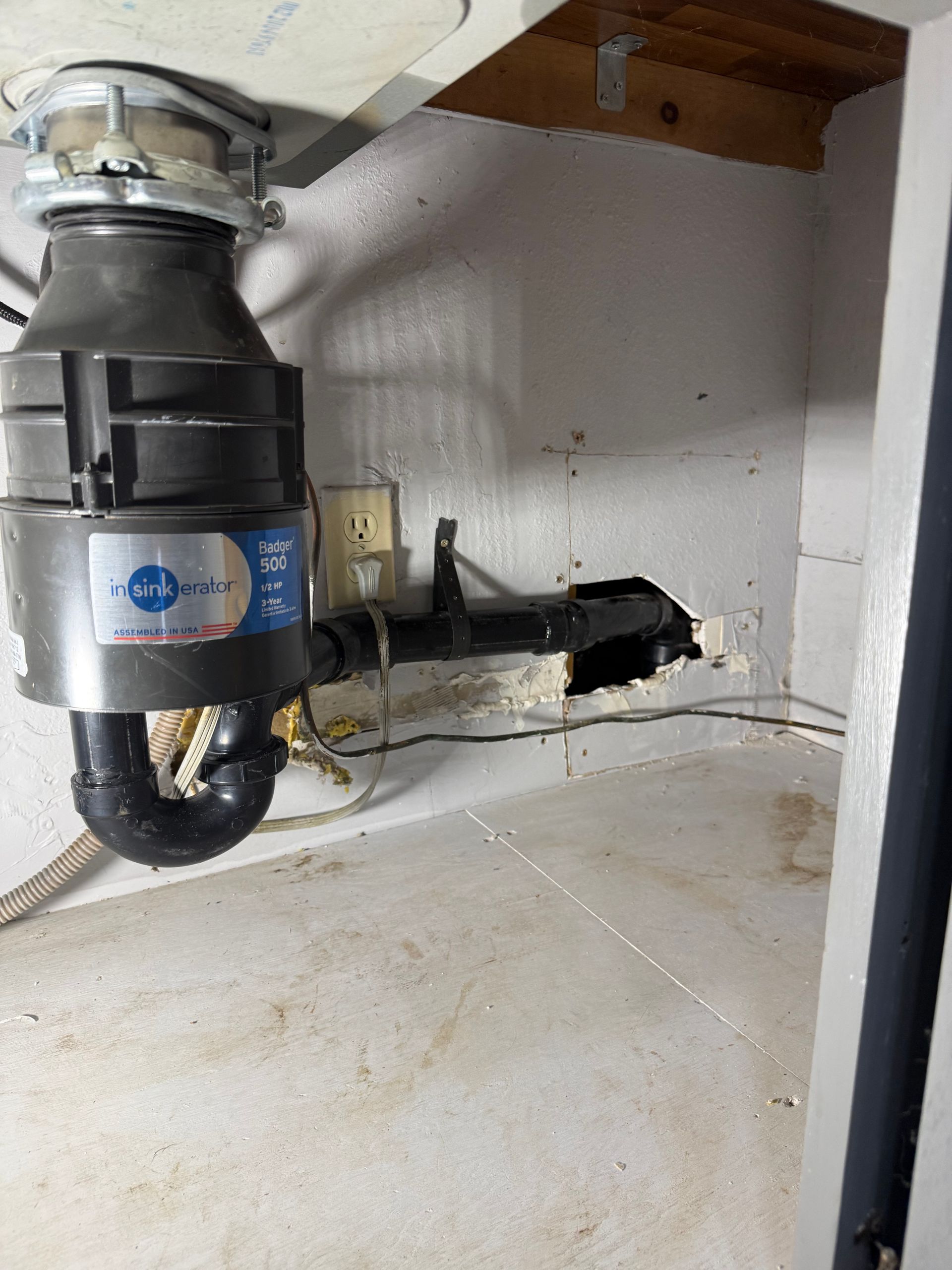 A garbage disposal connected to a black drain pipe in a cabinet under a kitchen sink with a wall opening nearby.